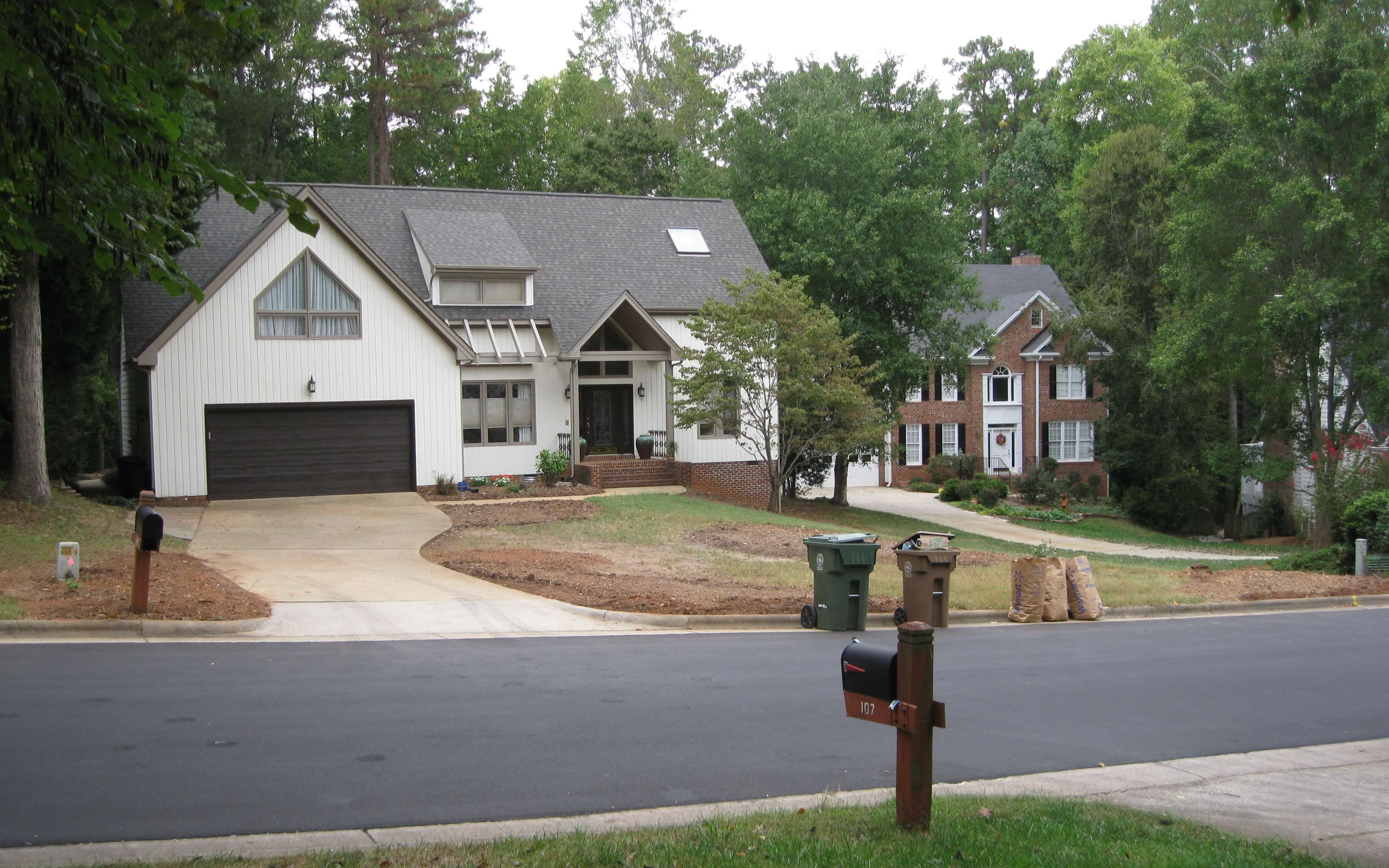 Suburban street view showing two houses with driveways, mailboxes, trash bins, and trees in the background.