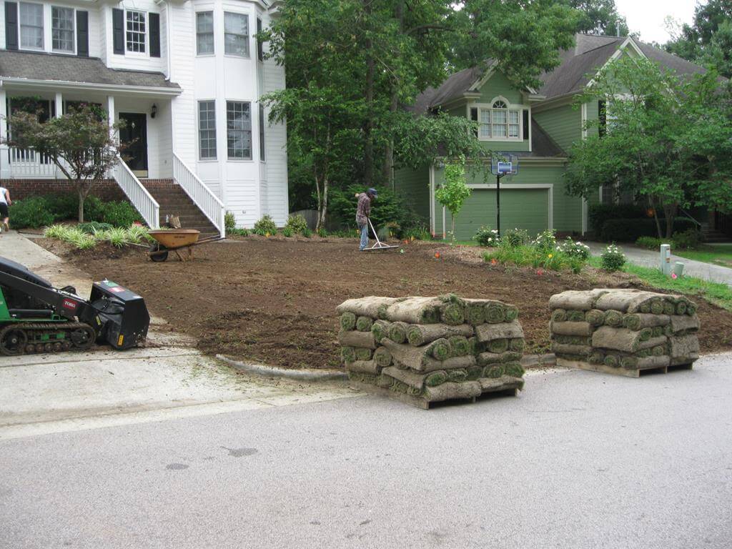 Backyard lawn renovation with two stacks of rolled sod on pallets and a man preparing soil in front of a white house and a green house.