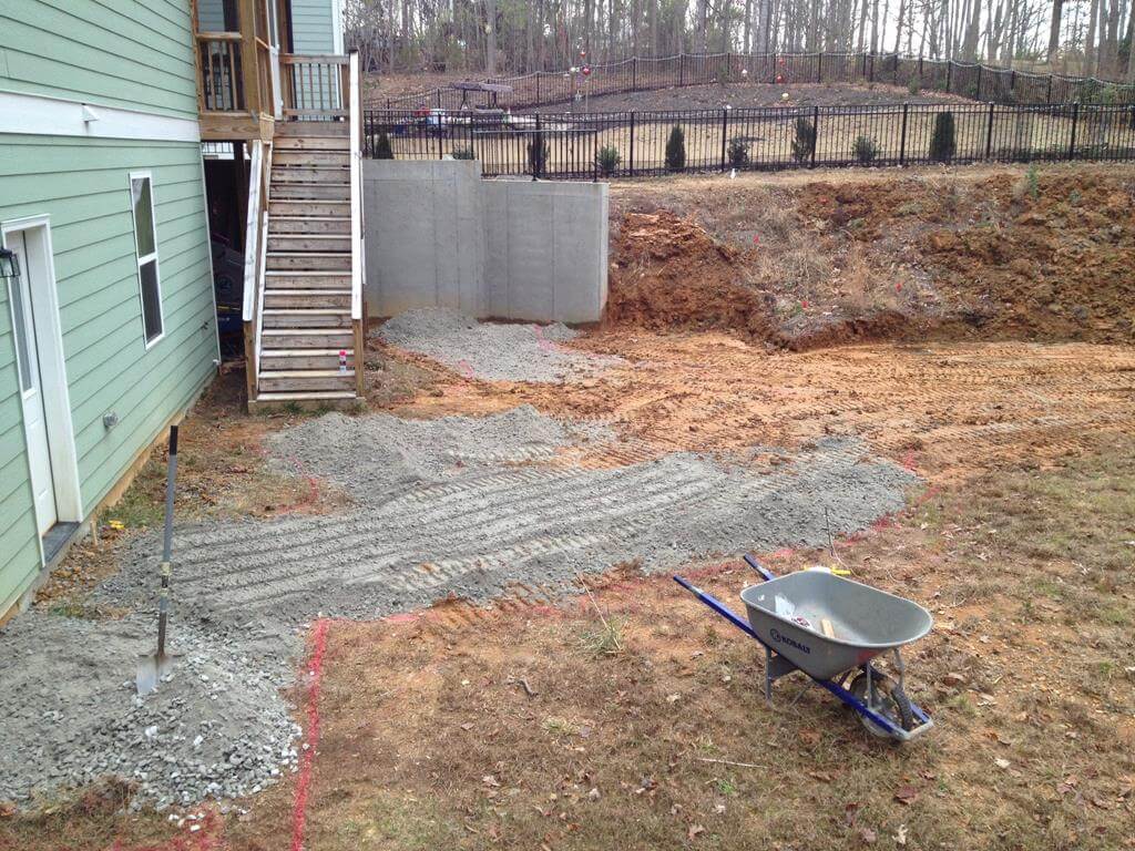 Yard under construction next to a green house with gravel spread on the ground, a wheelbarrow, and a wooden staircase.