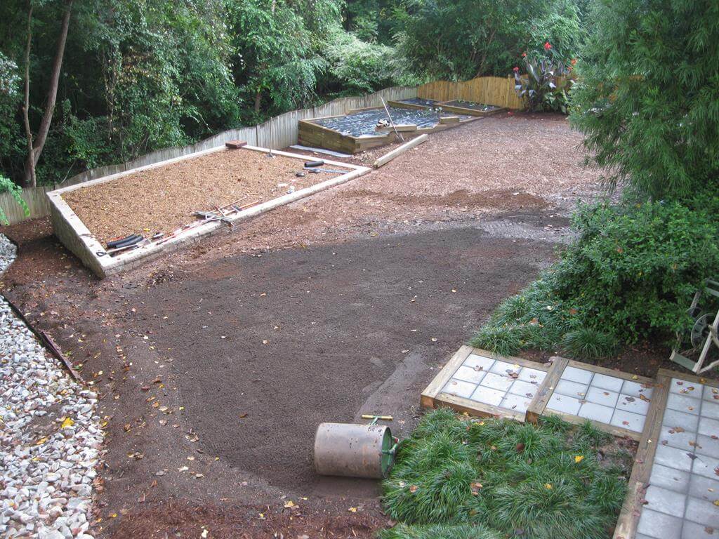 Backyard garden under construction with raised planting beds filled with soil and mulch, surrounded by trees and shrubs.