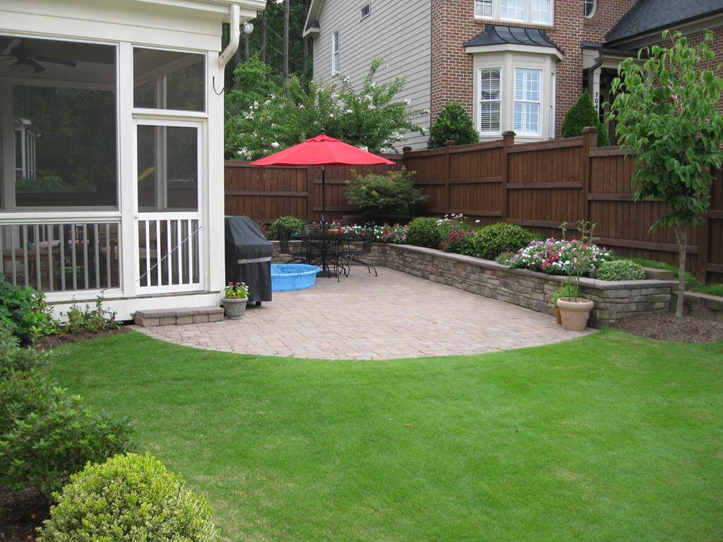 Backyard patio with stone pavers, black metal table and chairs, red umbrella, small blue kiddie pool, and raised stone flower bed along wooden fence.