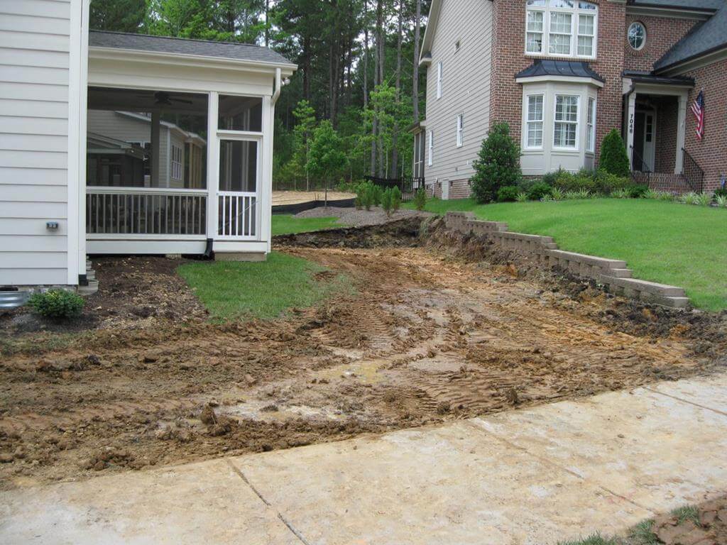 Backyard area between two houses showing a partially cleared and dug-up dirt yard with tire tracks, beside a green lawn and a small retaining wall.