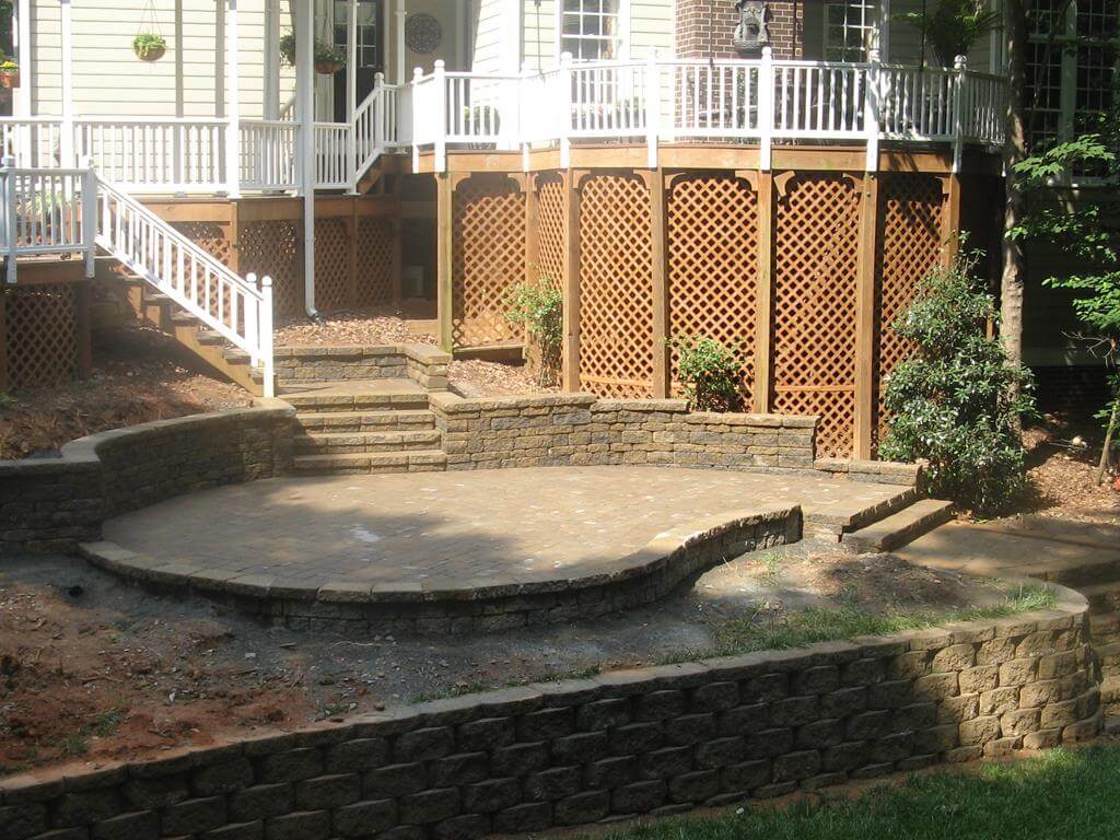 Backyard patio with a curved stone retaining wall, multi-level stone steps, and a wooden deck with white railings above.