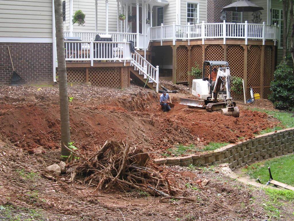 Backyard excavation near a raised wooden deck with a man sitting in the dirt and an excavator nearby.