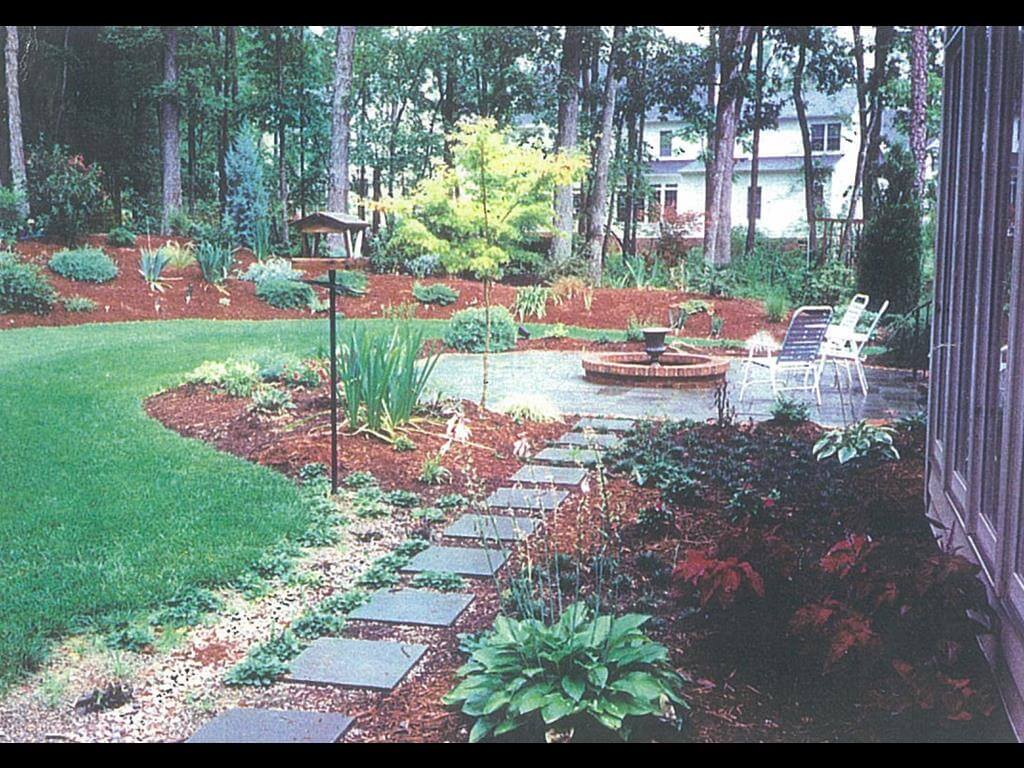 Backyard garden with a stone pathway, bird feeder, green lawn, various plants, trees, and a patio with chairs and a circular fountain.