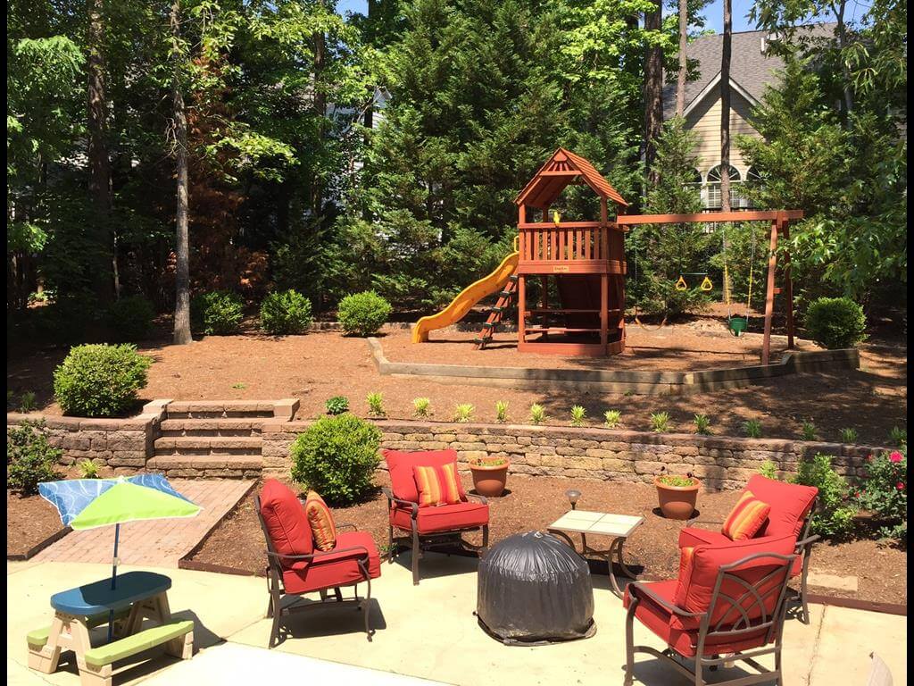 Backyard patio with red cushioned chairs around a covered fire pit and a wooden playset with a slide and swings in the background.