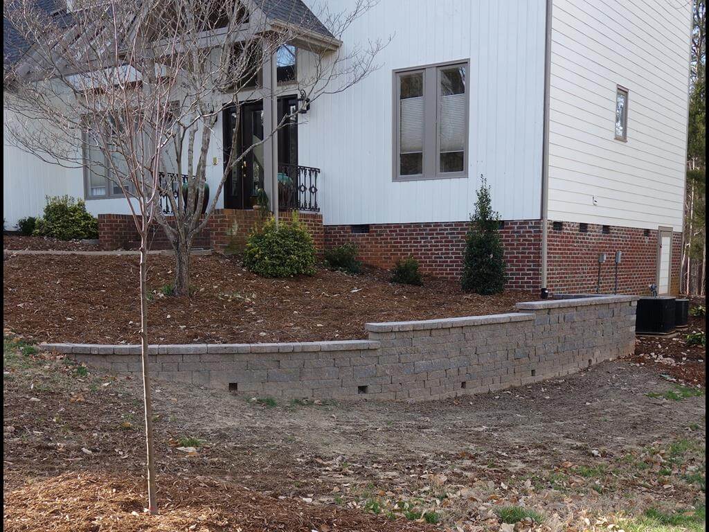 Backyard of a house with a tiered retaining wall made of gray bricks, mulched landscaping, and leafless trees.