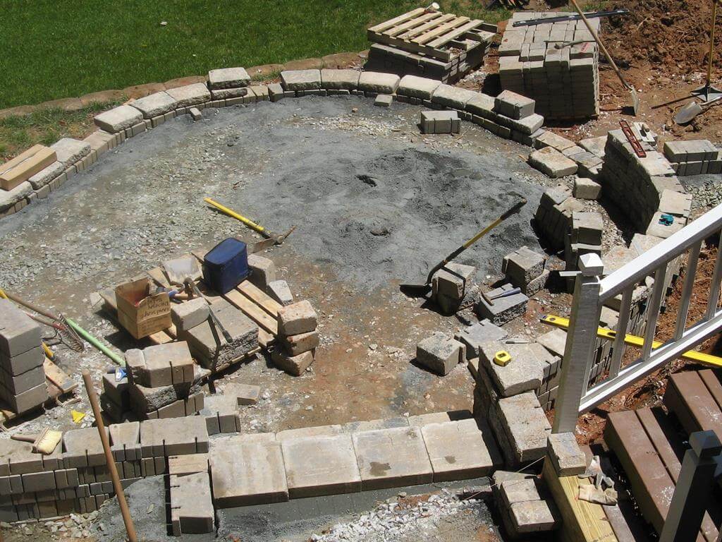 Construction site showing circular base being prepared with stacked pavers and landscaping tools around.