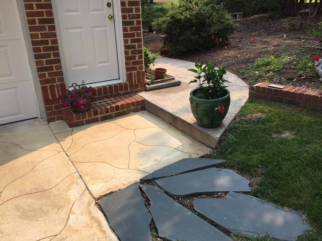 Cracked stone stepping path leading to a raised concrete walkway with a green potted plant near a brick house entrance.