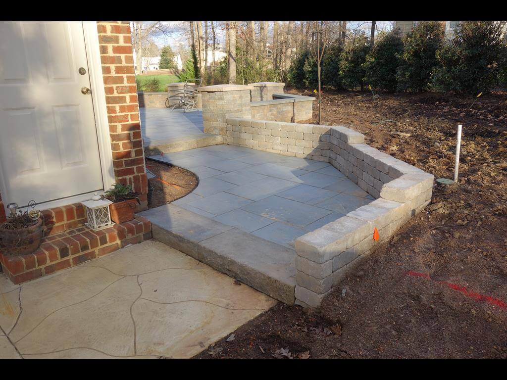 Backyard patio area with stone tile flooring, surrounding low retaining walls, and adjacent door with brick steps.