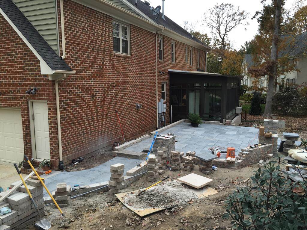 Backyard patio under construction with gray stone tiles being laid, stacks of bricks, and construction tools near a brick house.