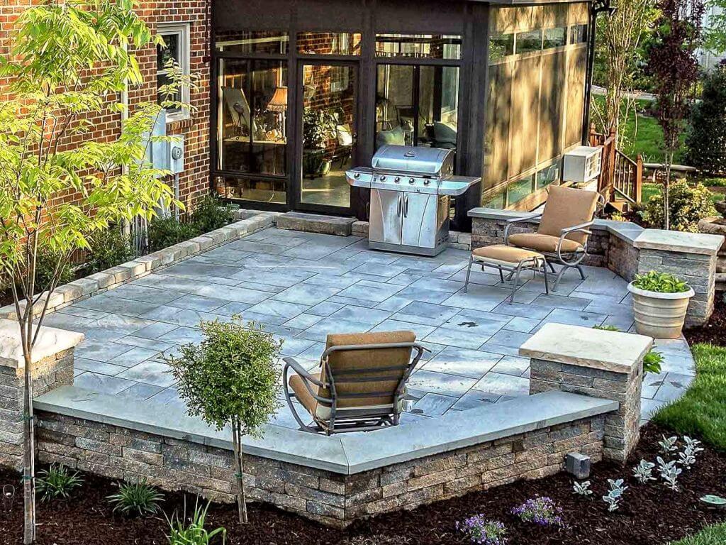 Stone patio with two cushioned chairs, a stainless steel grill, and surrounding greenery outside a brick house.