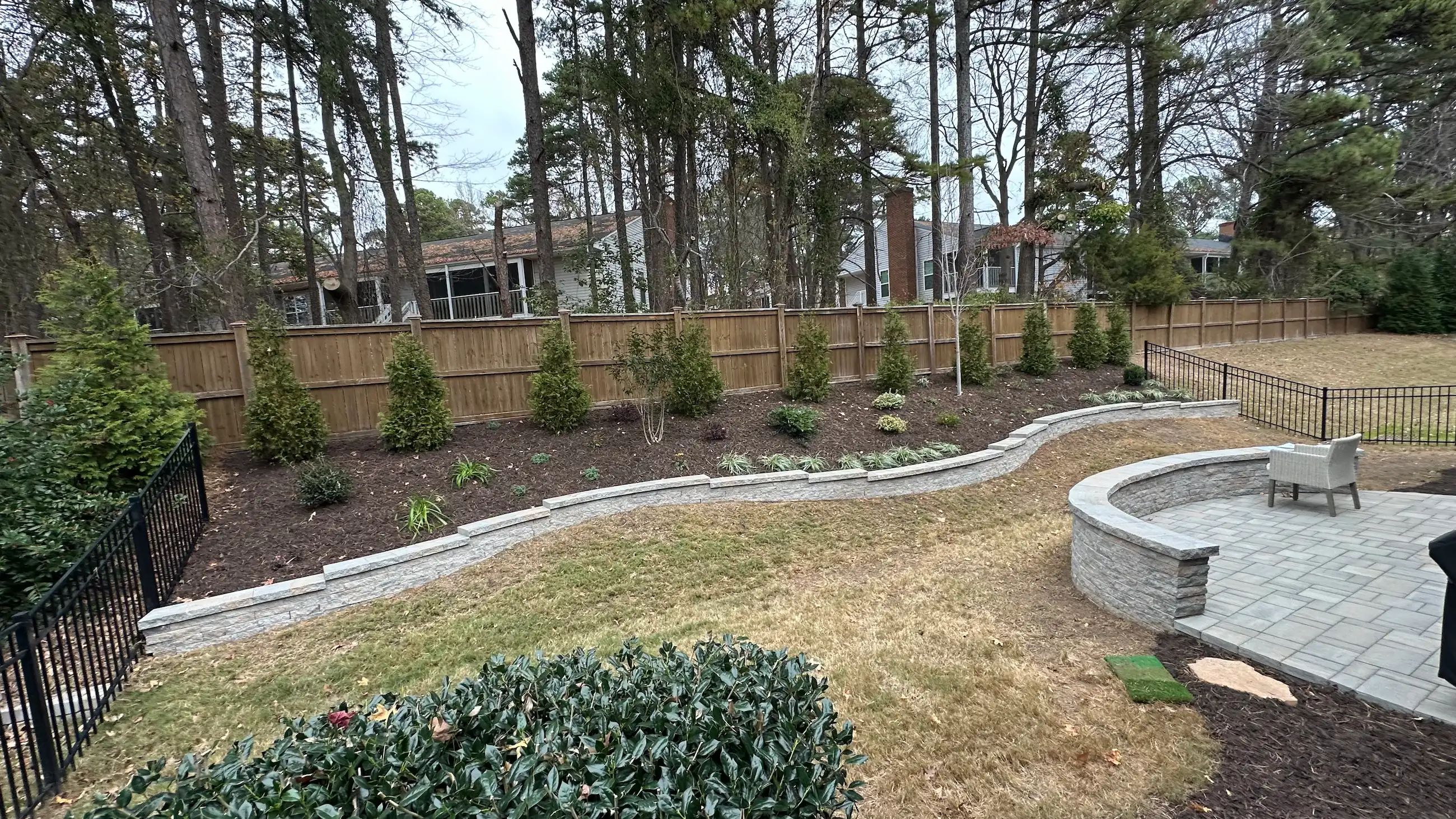 A landscaped yard featuring a stone retaining wall, flower beds with colorful blooms, and shrubs, alongside a modern white house with a sloped roof.