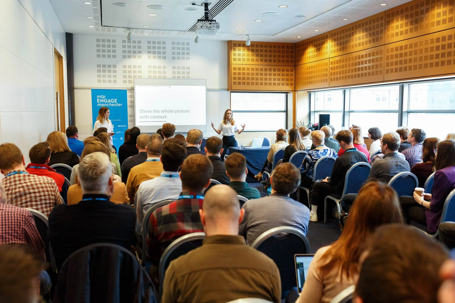 Speaker presenting to a seated audience in a conference room with a projector and large windows.