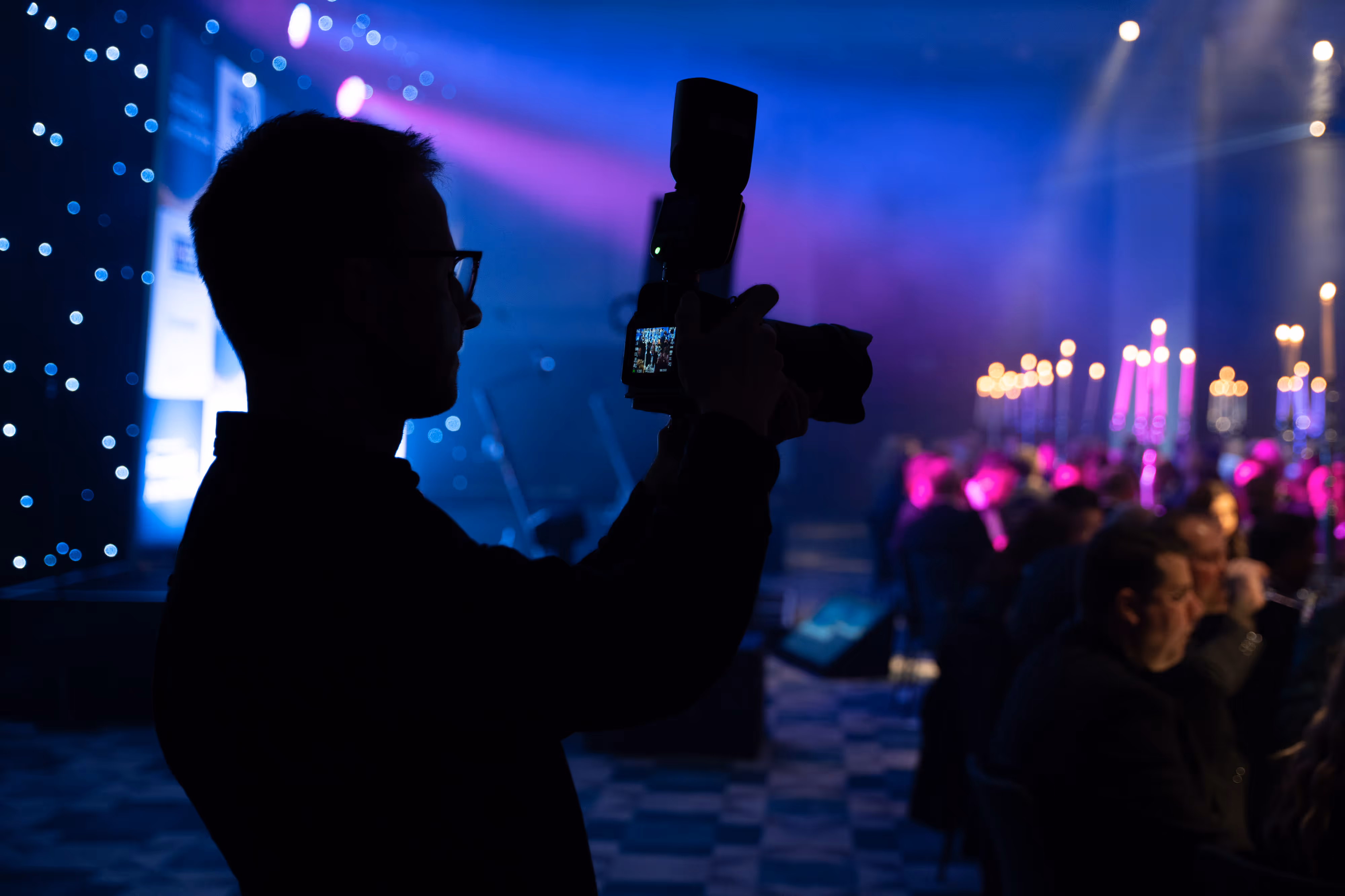 Silhouette of a photographer holding a camera with flash in a dimly lit event room with purple and blue lighting and seated guests.