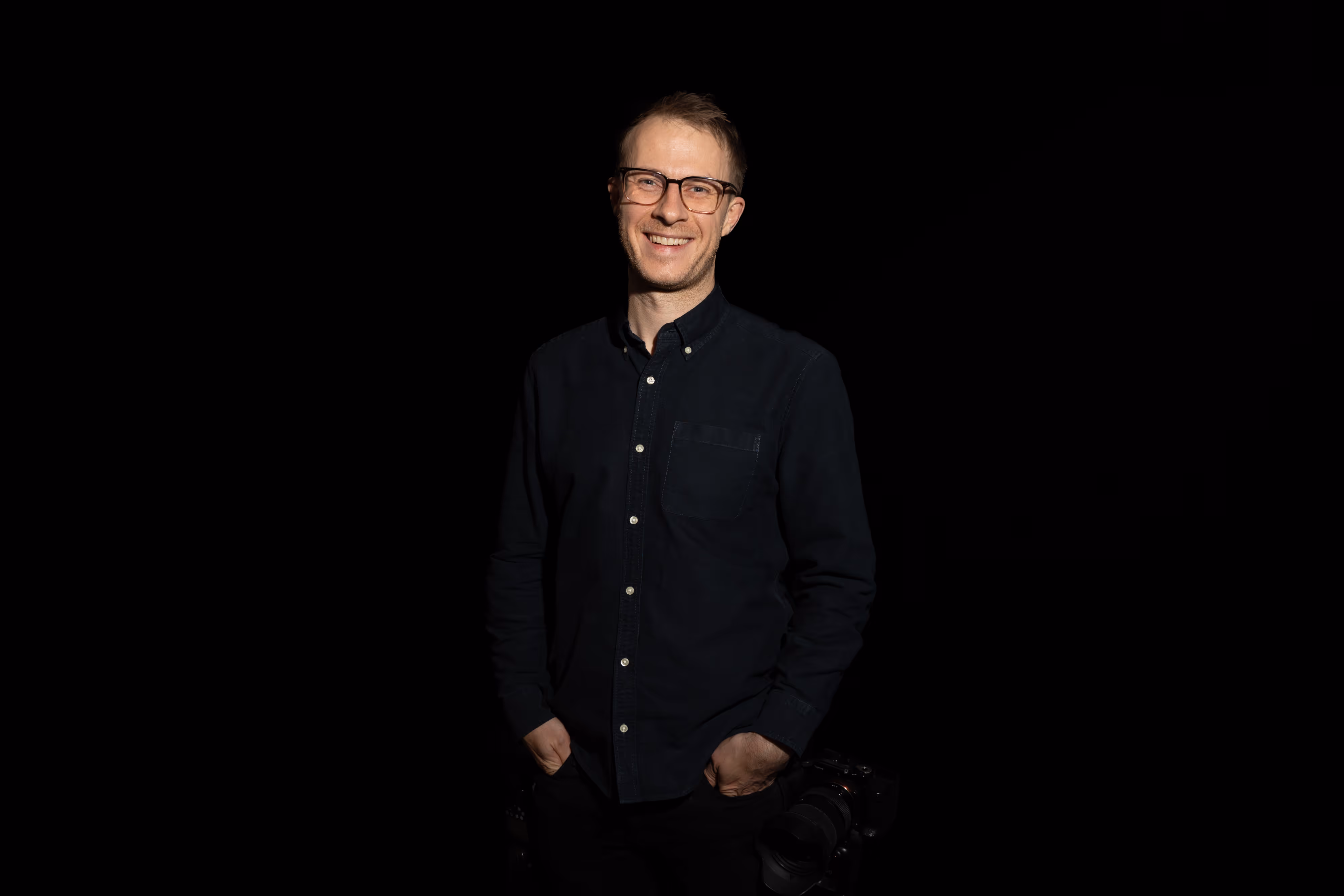 Smiling man wearing glasses and a black button-up shirt standing against a black background with a camera hanging by his side.
