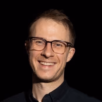 Smiling man (Alex Mead) wearing glasses against a black background.