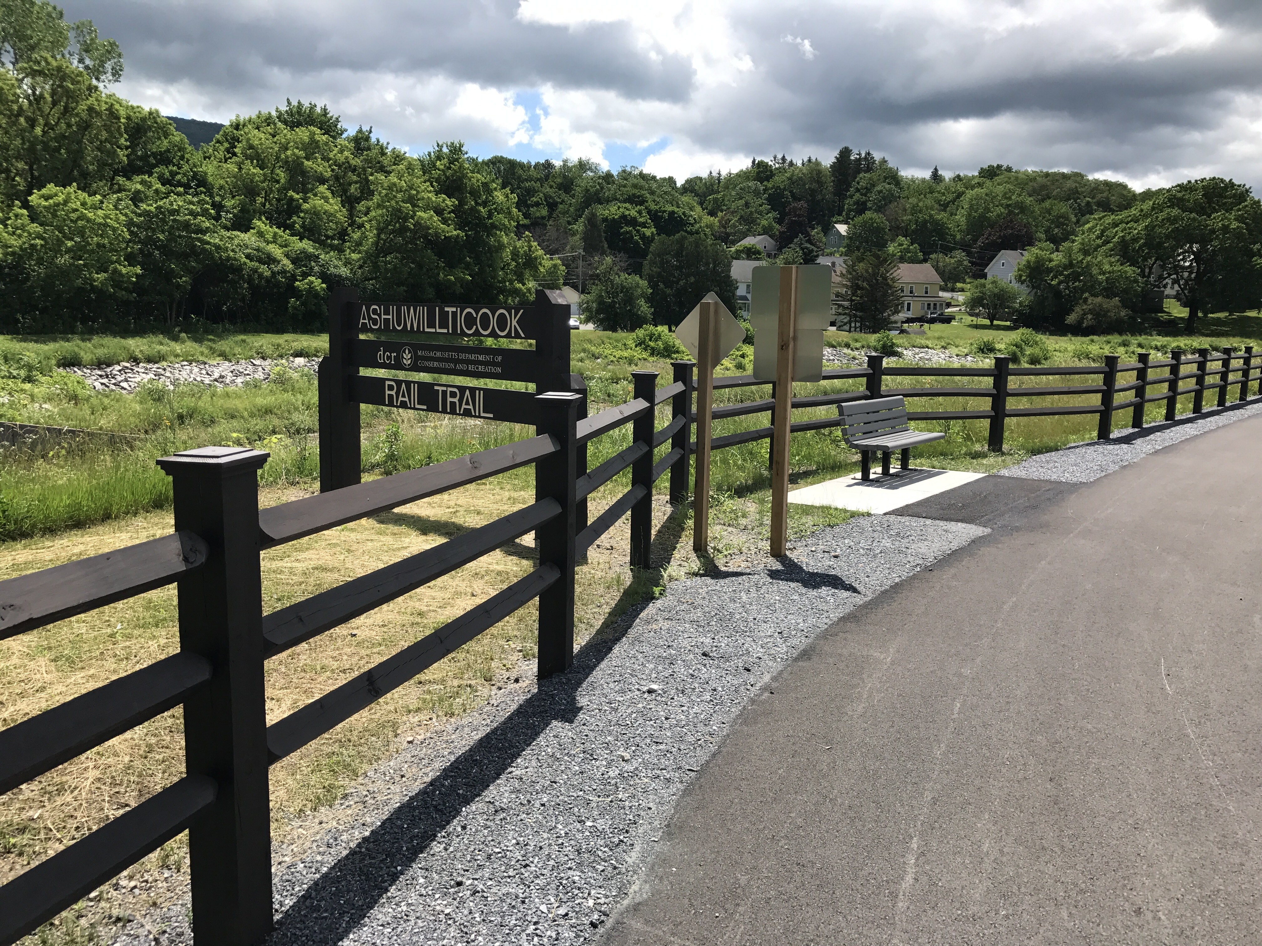 Gray clouds hover over dark, leafy green trees. In the midground, a dark colored fence runs along the asphalt path. On the left, a sign with the words Ashuwillticook Rail Trail is outside the fence. On the right, a bench faces the path inside of the fence.