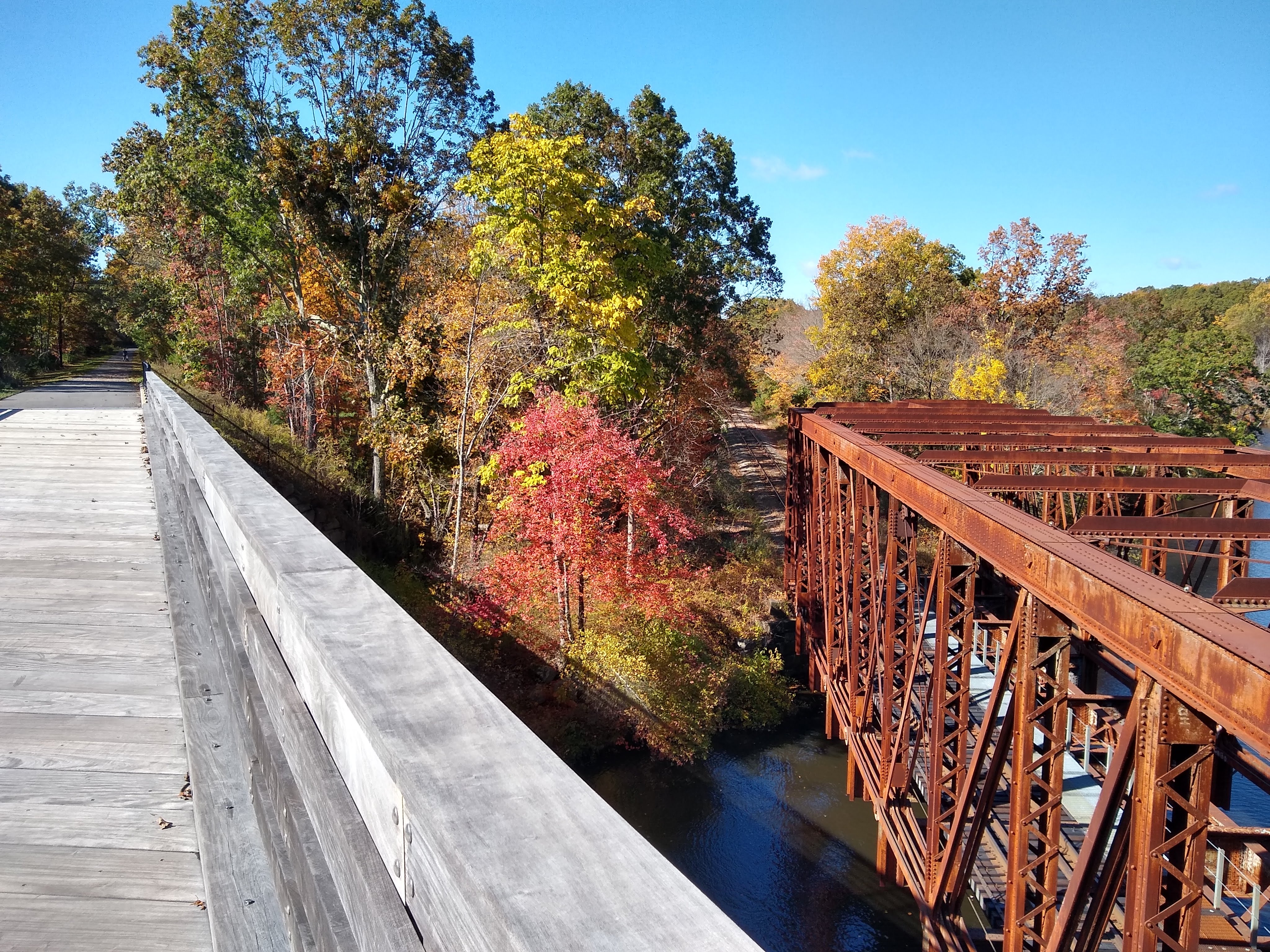 A brightly colored photograph features a light-gray wood-decked bridge connecting to the asphalt trail in the distance on the left; red, orange, yellow, and green-leafy trees in the middle; and a rust-colored steel truss bridge over a waterway on the right