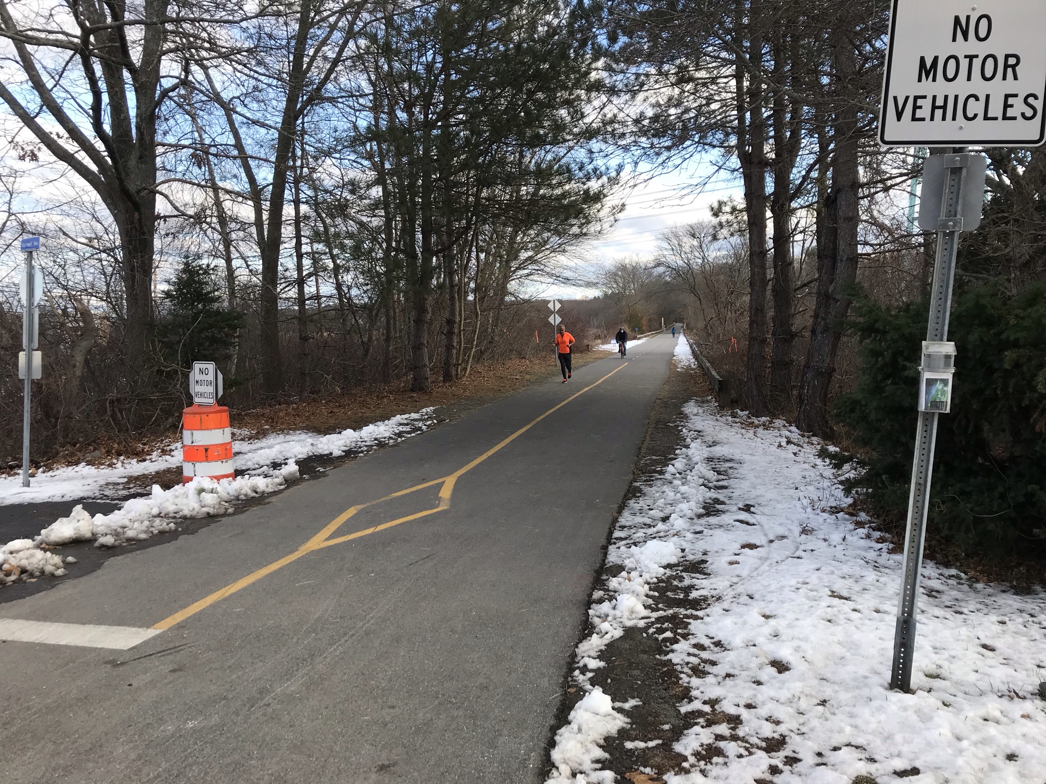 Some snow flanks the asphalt trail on the right and left sides of the image. A yellow solid centerline runs down the asphalt path in the middle of the image. Three people are visible on the path, either jogging or using a bicycle. 