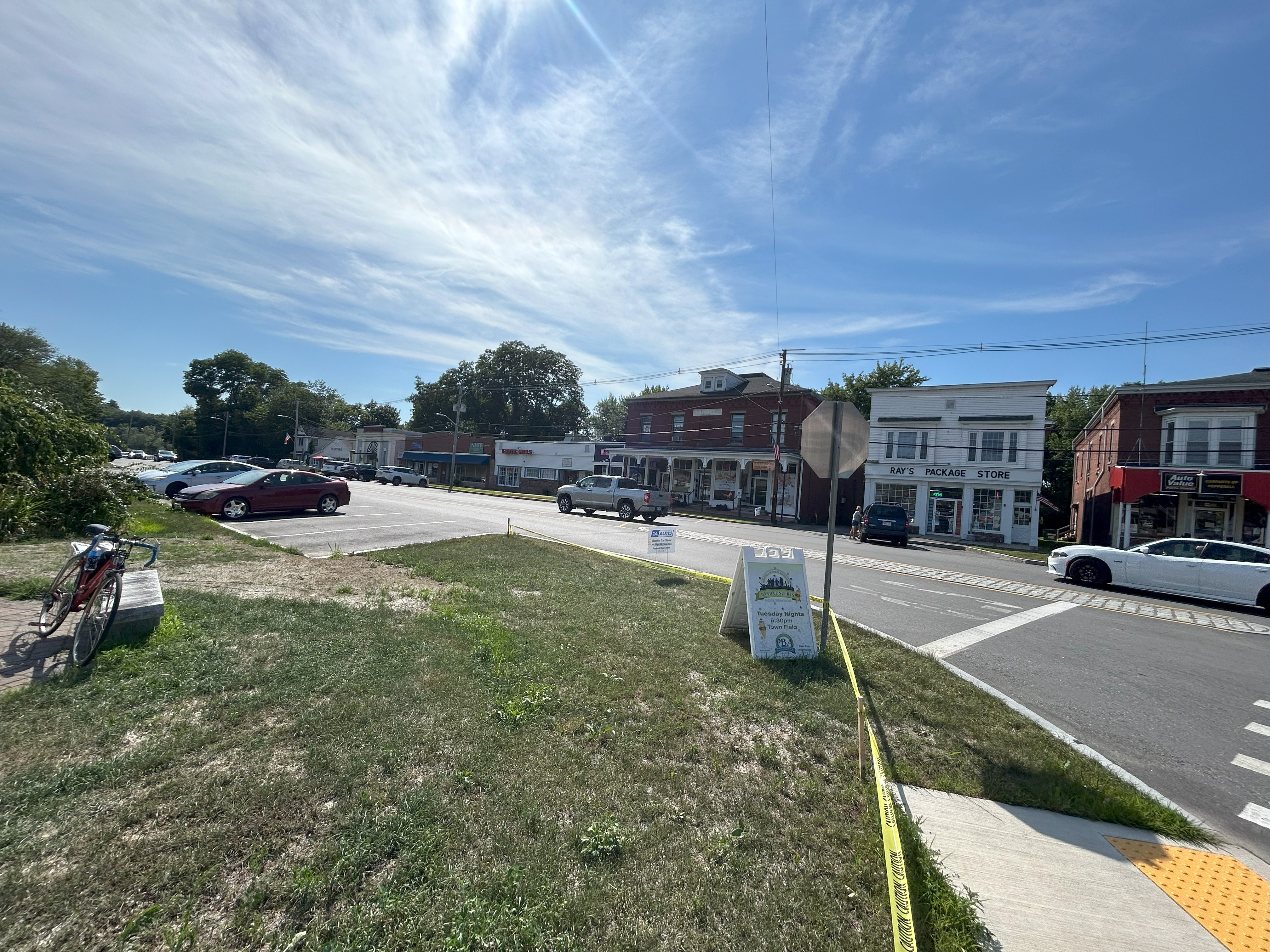 A few one- and two-story buildings sit in the background along a road with a few cars. A bicycle is visible on the left, and a yellow tactile warning strip at a curb cut leading to a crosswalk is just visible in the right lower corner.