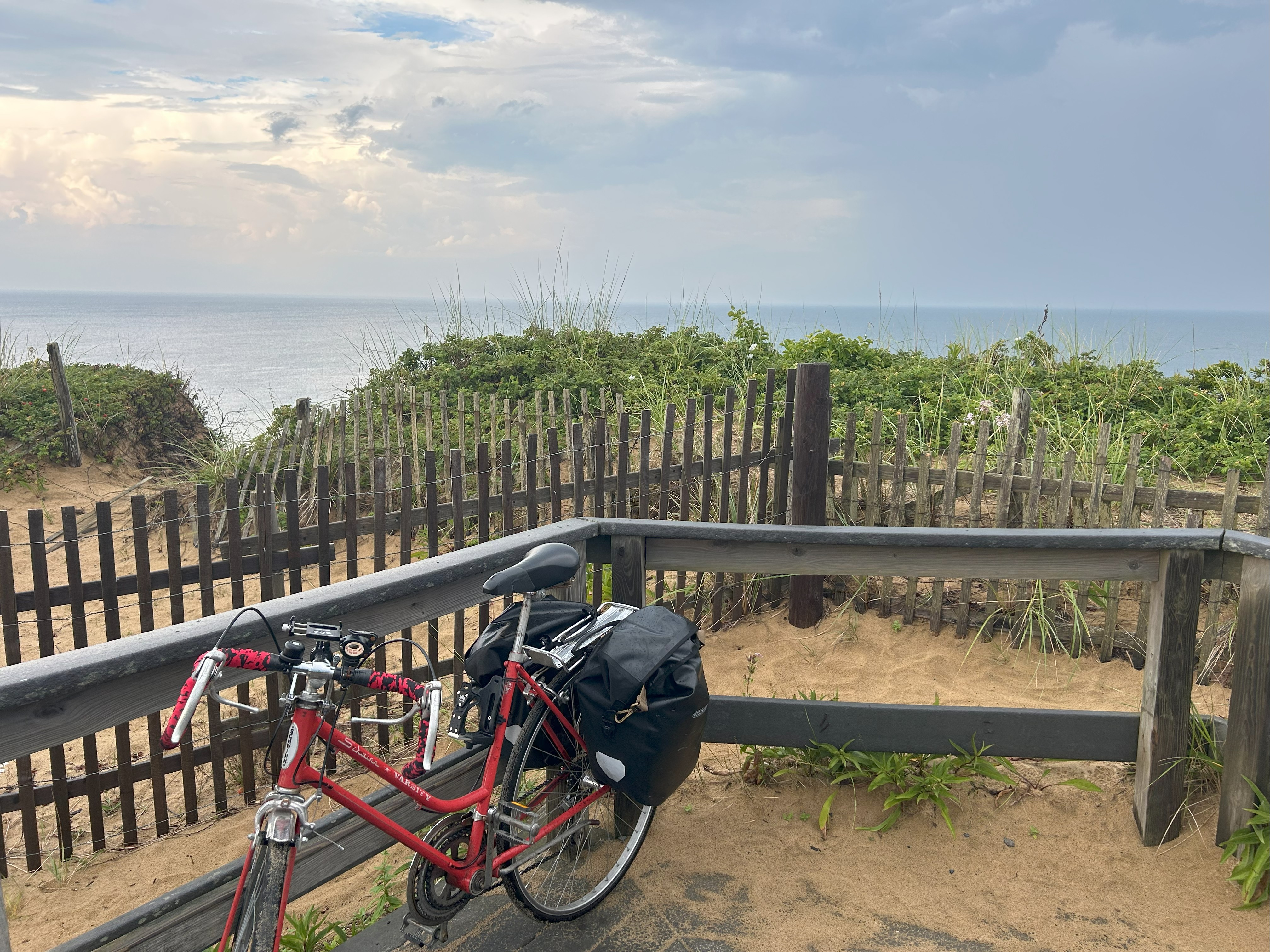 A cloudy sky at sunset hovers over the ocean in the background of the image. In the midground, low green shrubs and a sandy embankment lead to two wooden fences and a red bicycle in the foreground. 