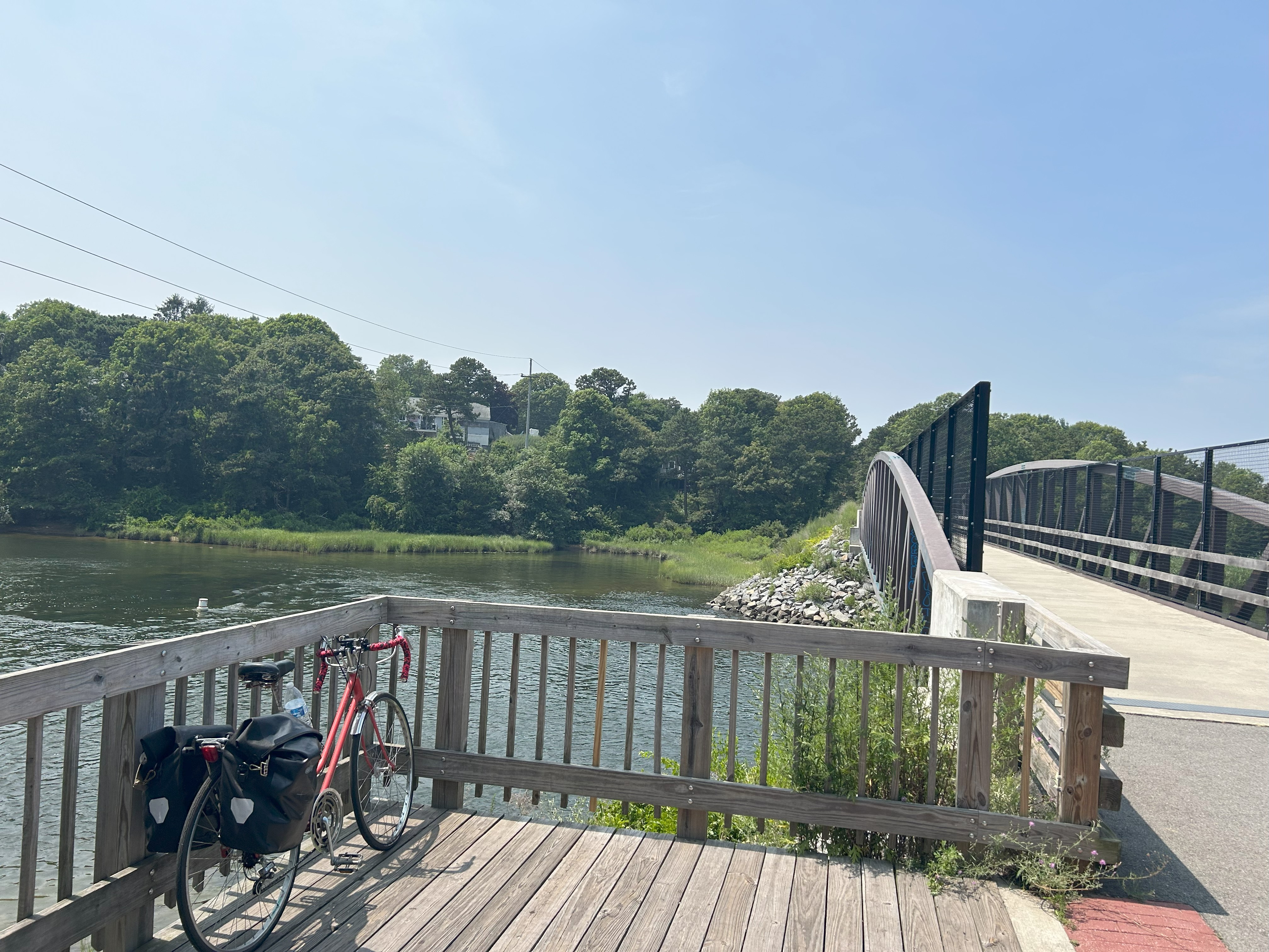 A small wooden overlook on the left side of the image has a red bicycle facing the Bass River. On the right side of the image, a light-colored path on a bridge with a black fence crosses the Bass River toward the background of the image. 