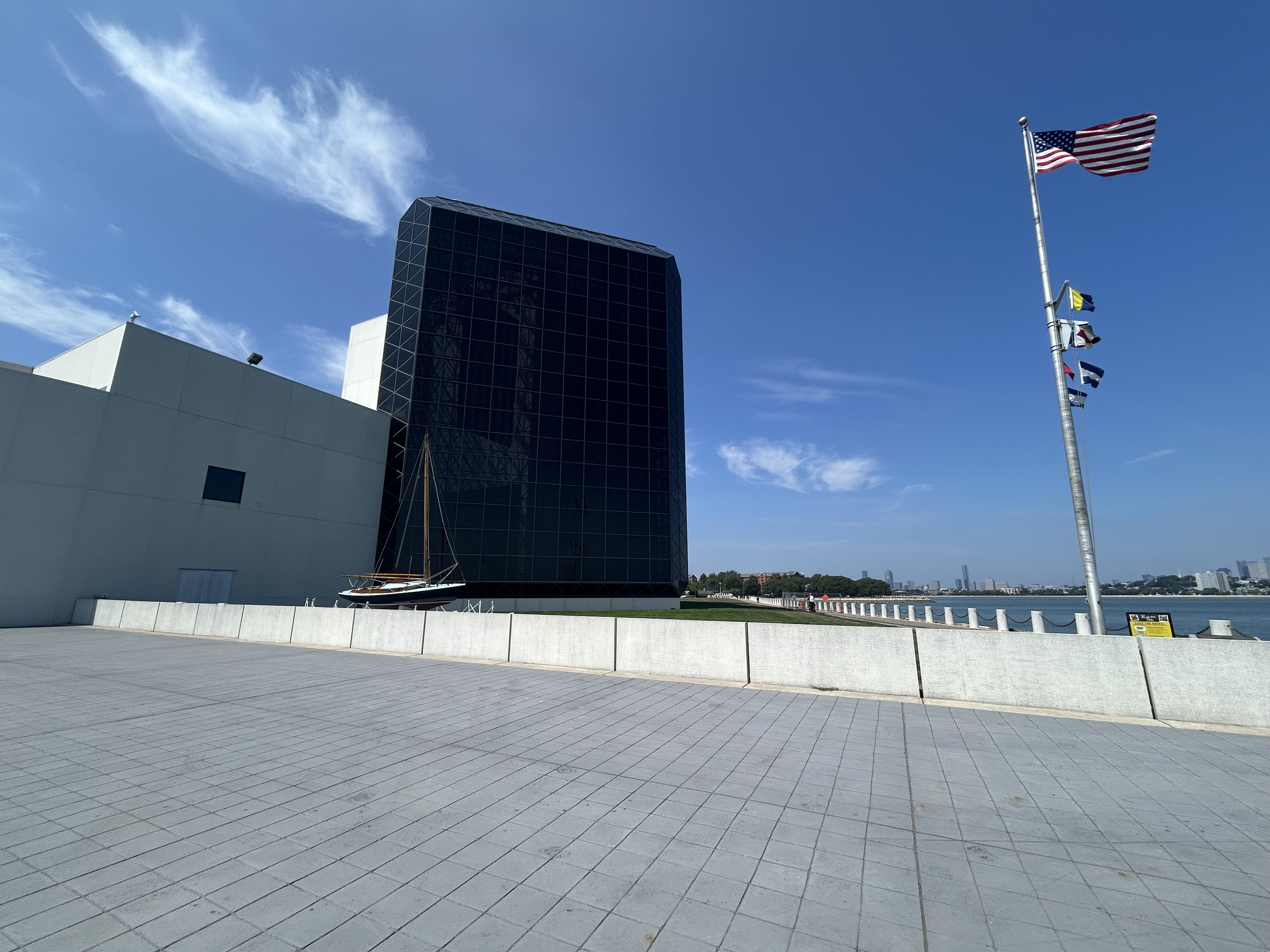  A large black glass building is in the center of the photo, connected to a smaller light-gray building on its left. In front of the buildings is a sailboat. On the right side of the image is a flagpole flying a United States flag as well as nautical flags