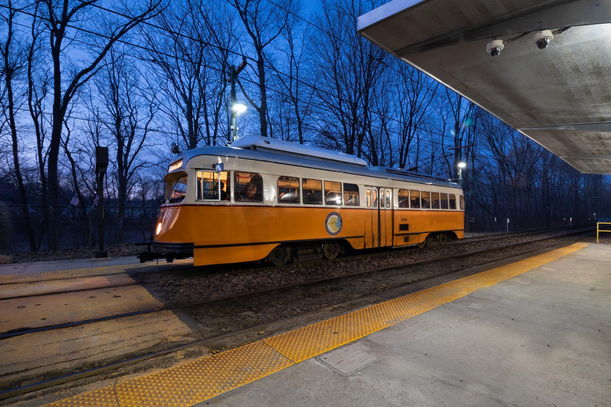 A single orange Mattapan trolley-car moves from the right side of the image to the left, under a night sky, with leafless trees in the background. 