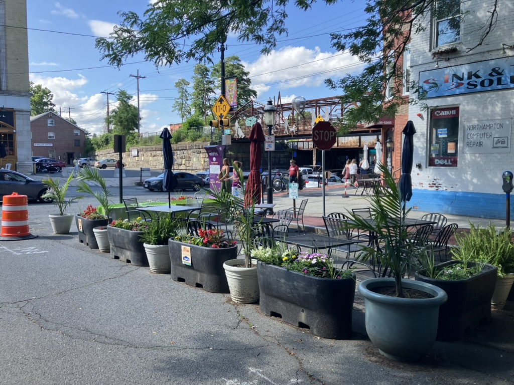 A bustling city intersection, featuring potted plants in the foreground, a small sidewalk cafe with tables and chairs in the midground, and a railroad bridge crossing over a street in the background. There are a handful of cars and people walking.