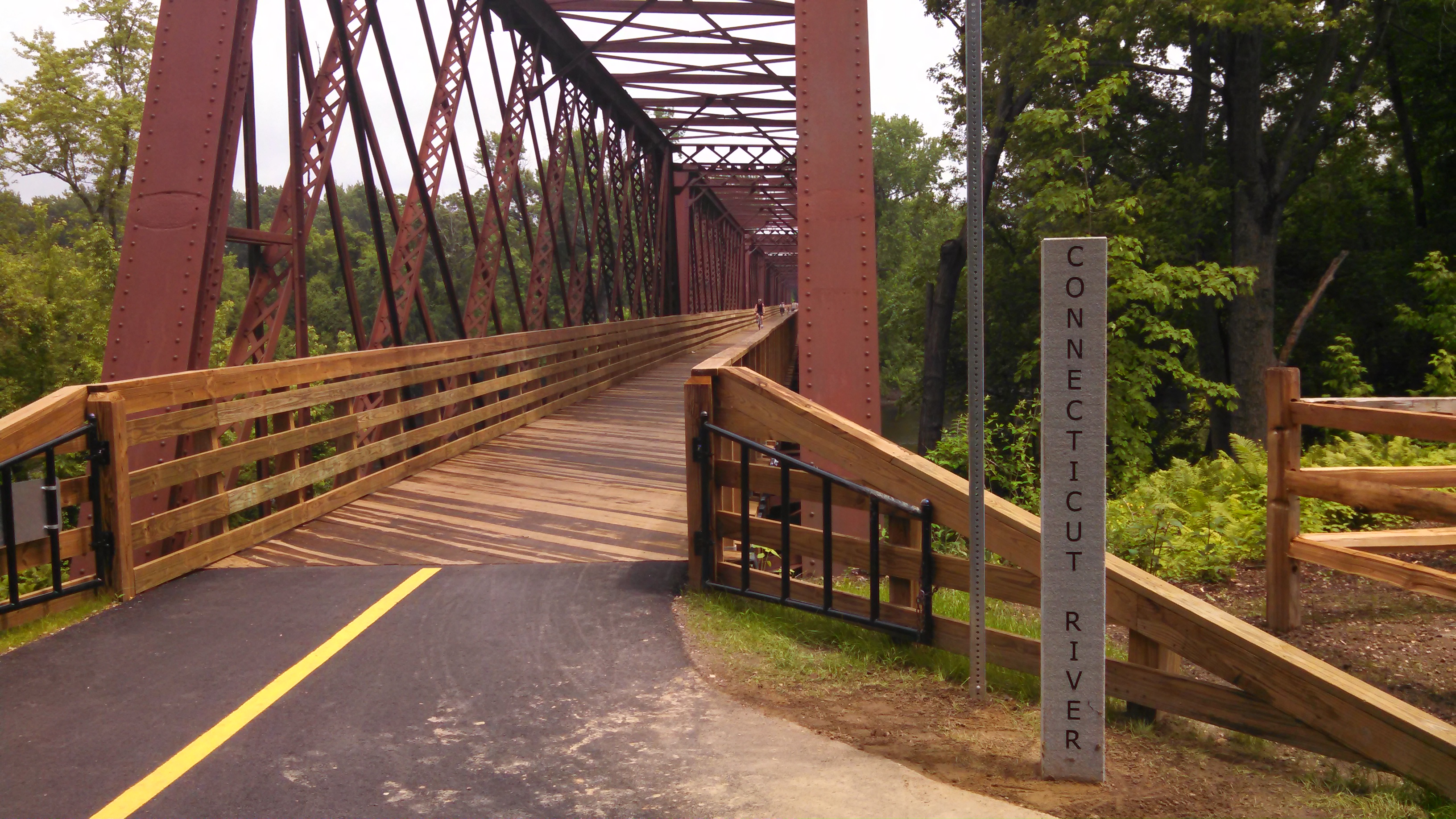A brownish red steel truss bridge with a wooden deck spans the left side of the image. Far in the distance, a person riding on the bridge on a bicycle is visible. To the right of the bridge is granite marker with the words Connecticut River.