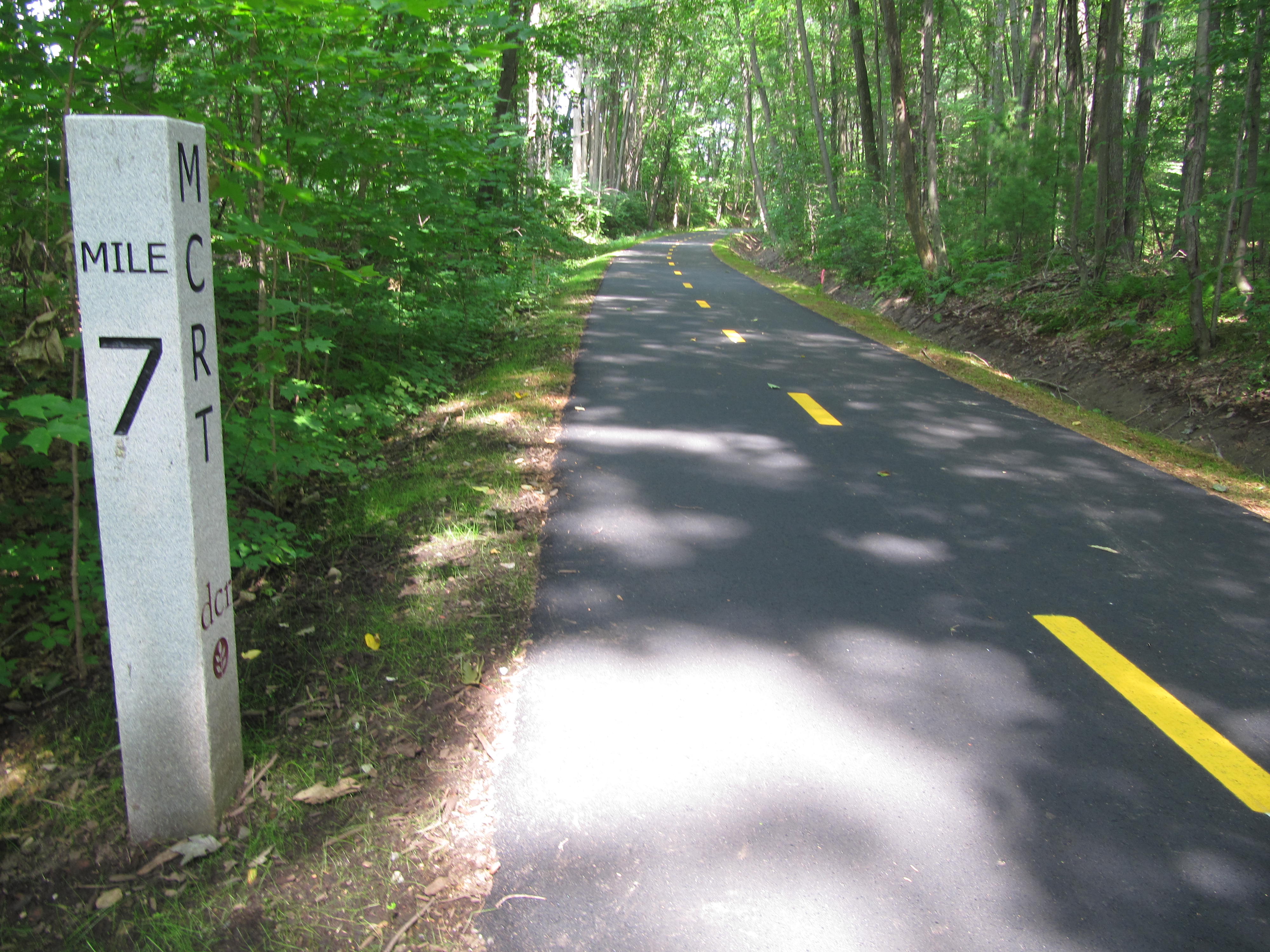 A granite mile marker post says “Mile 7” on one face and M C R T with the Department of Conservation and Recreation logo on another face. The post is on the left side of the image, next to the asphalt trail, which has a dotted yellow line.