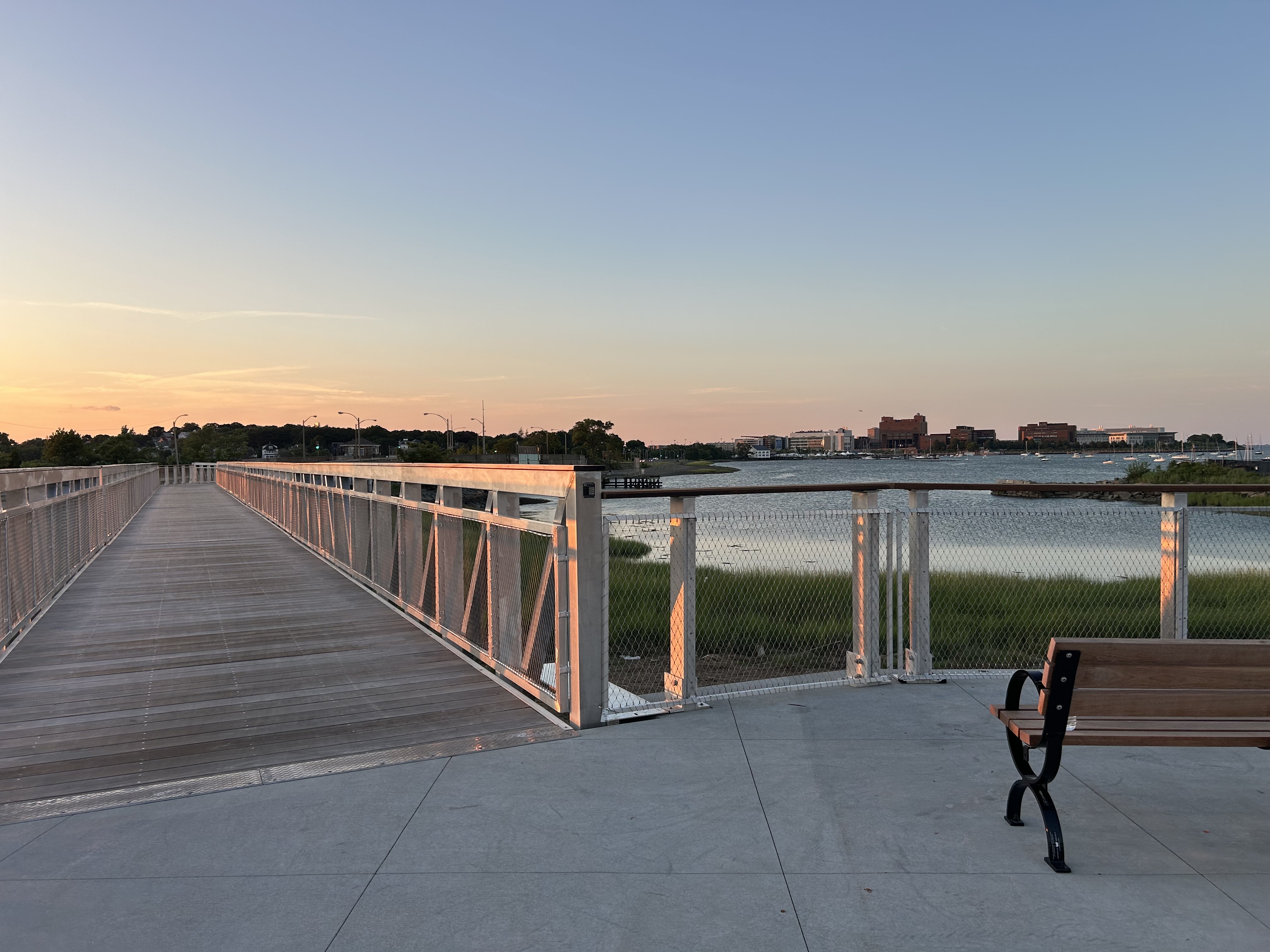 A light wood boardwalk with aluminum railings stretches from the foreground to the background of image, under a sunset sky, with water and a bench on the right side.
