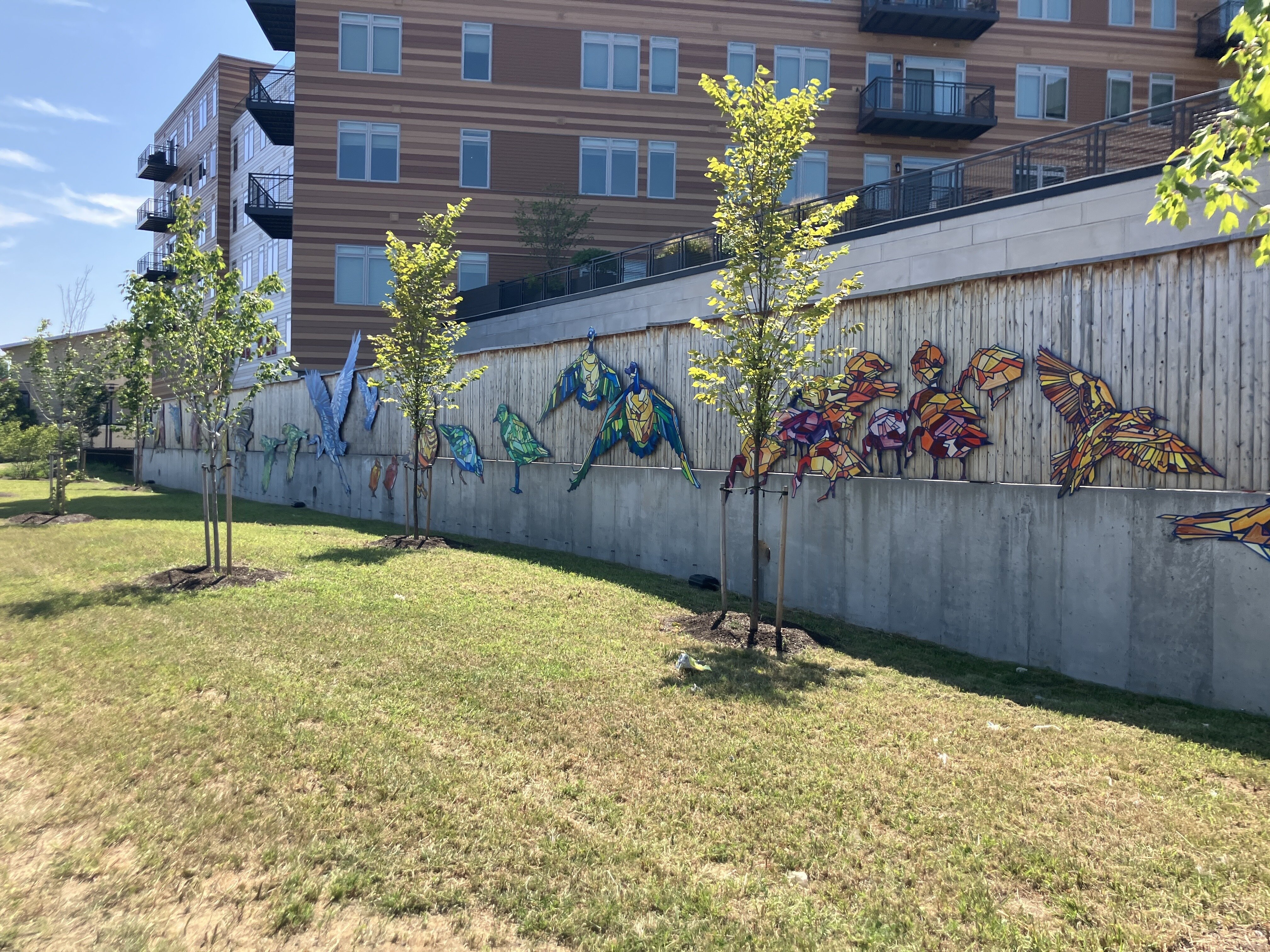 A strip of grass is on the left of the image, which leads to a wood and stone wall that features art of multicolored birds created to look like stained glass.
