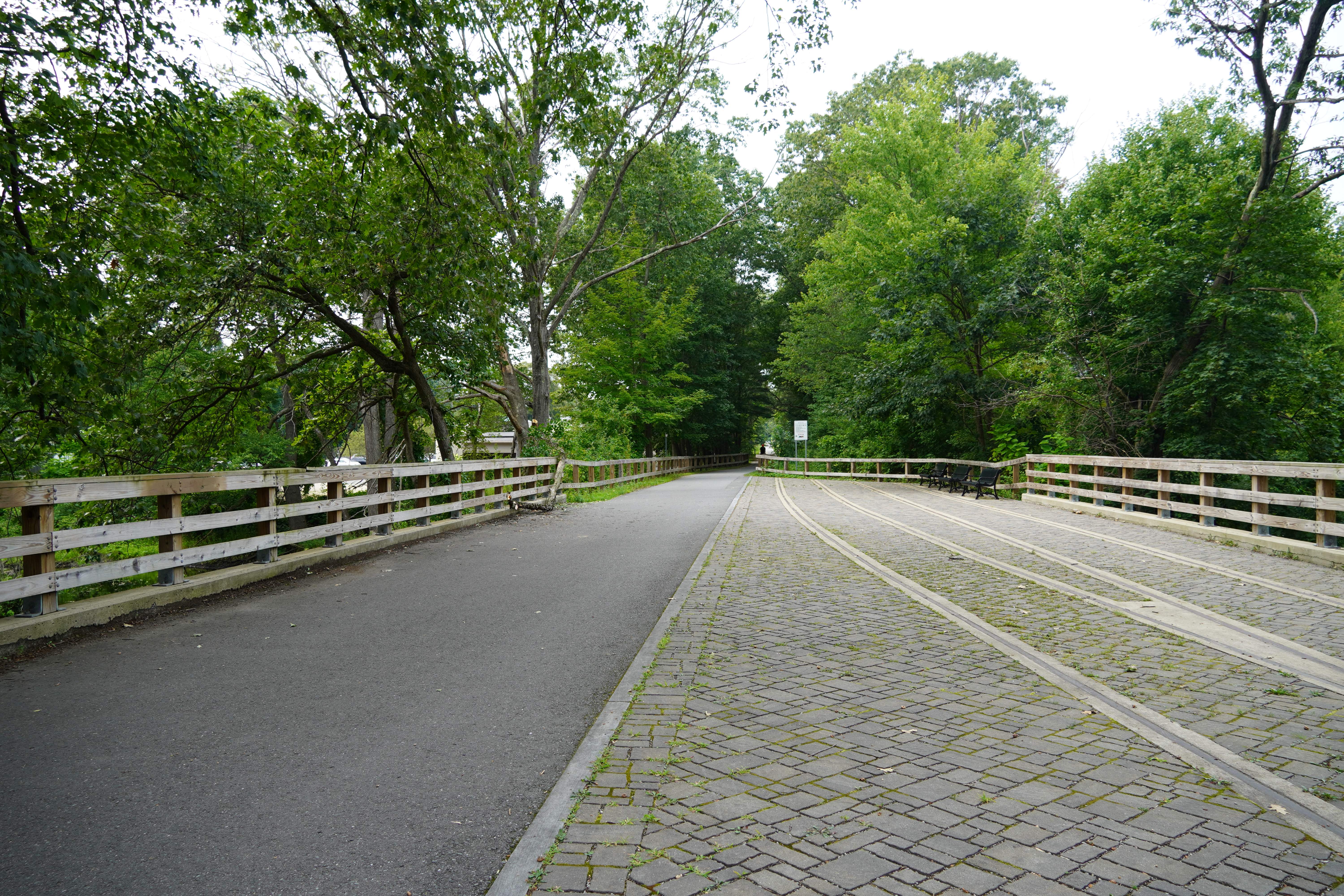 this picture shows the trail next to three benches for resting. theres also abandoned train tracks that have been surrounded by pavers to create a resting spot.