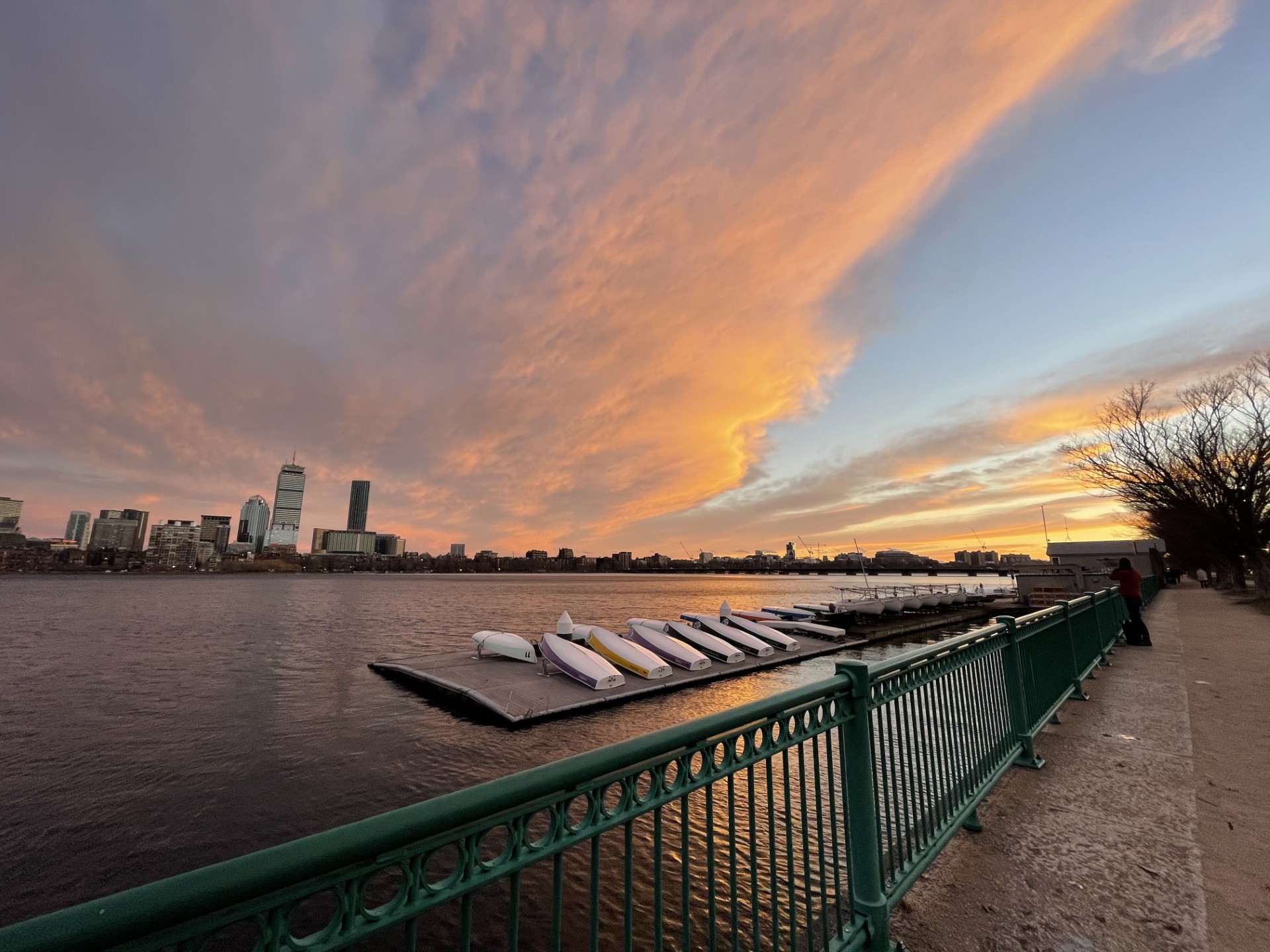 this photo shows the sky during sunset looking towards boston from the path's Cambridge side. there is a dock with upside down boats stored on it