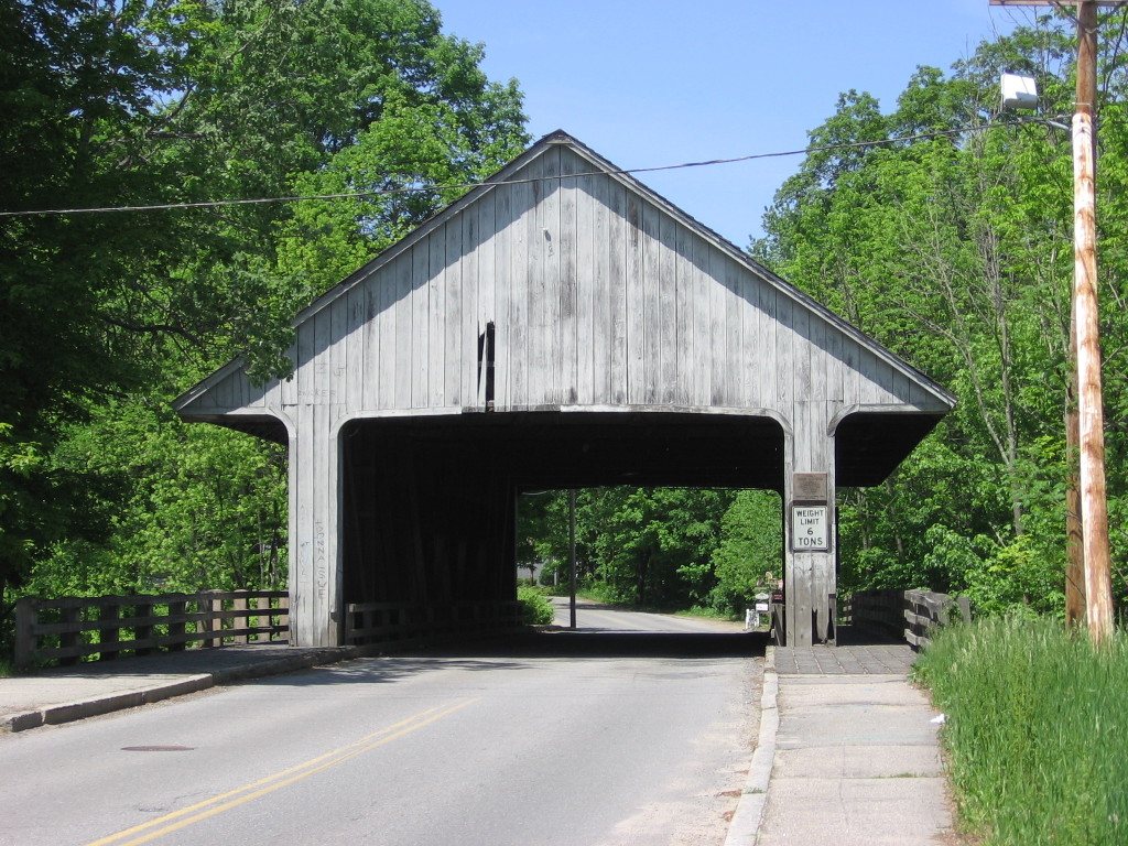A view of a wooden covered bridge with the road in the foreground. Trees are on either side