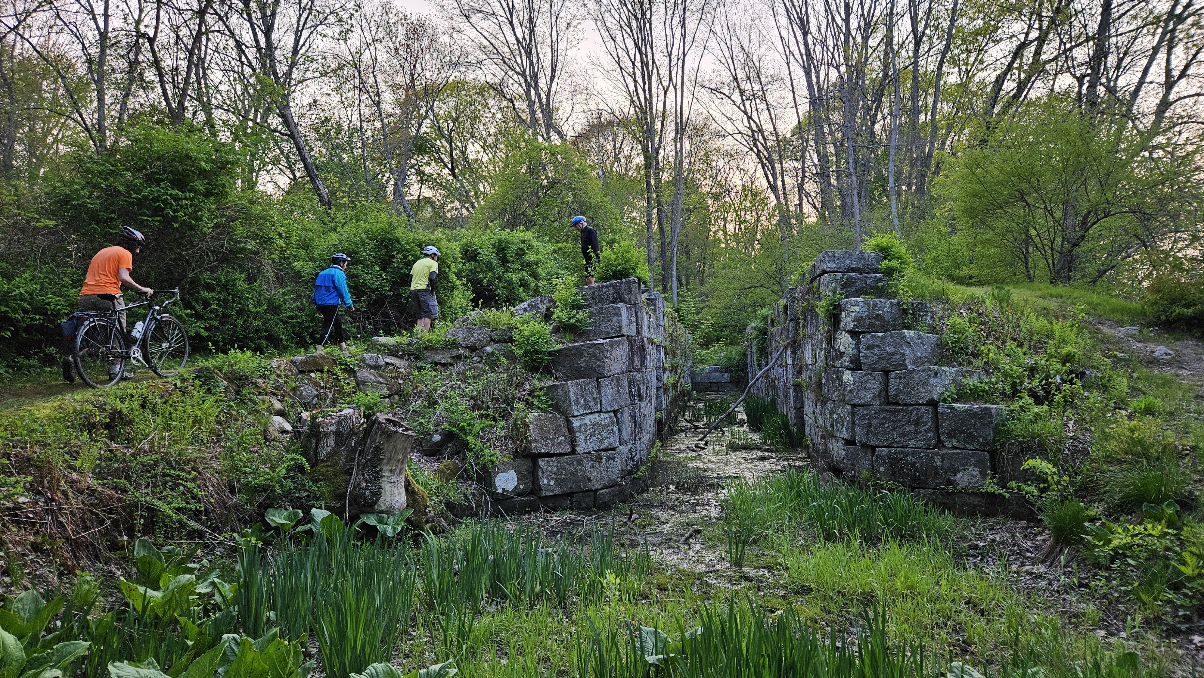 this photo shows the Millville lock in the woods, with three people climging up the left side with bikes. in the center is a small body of water