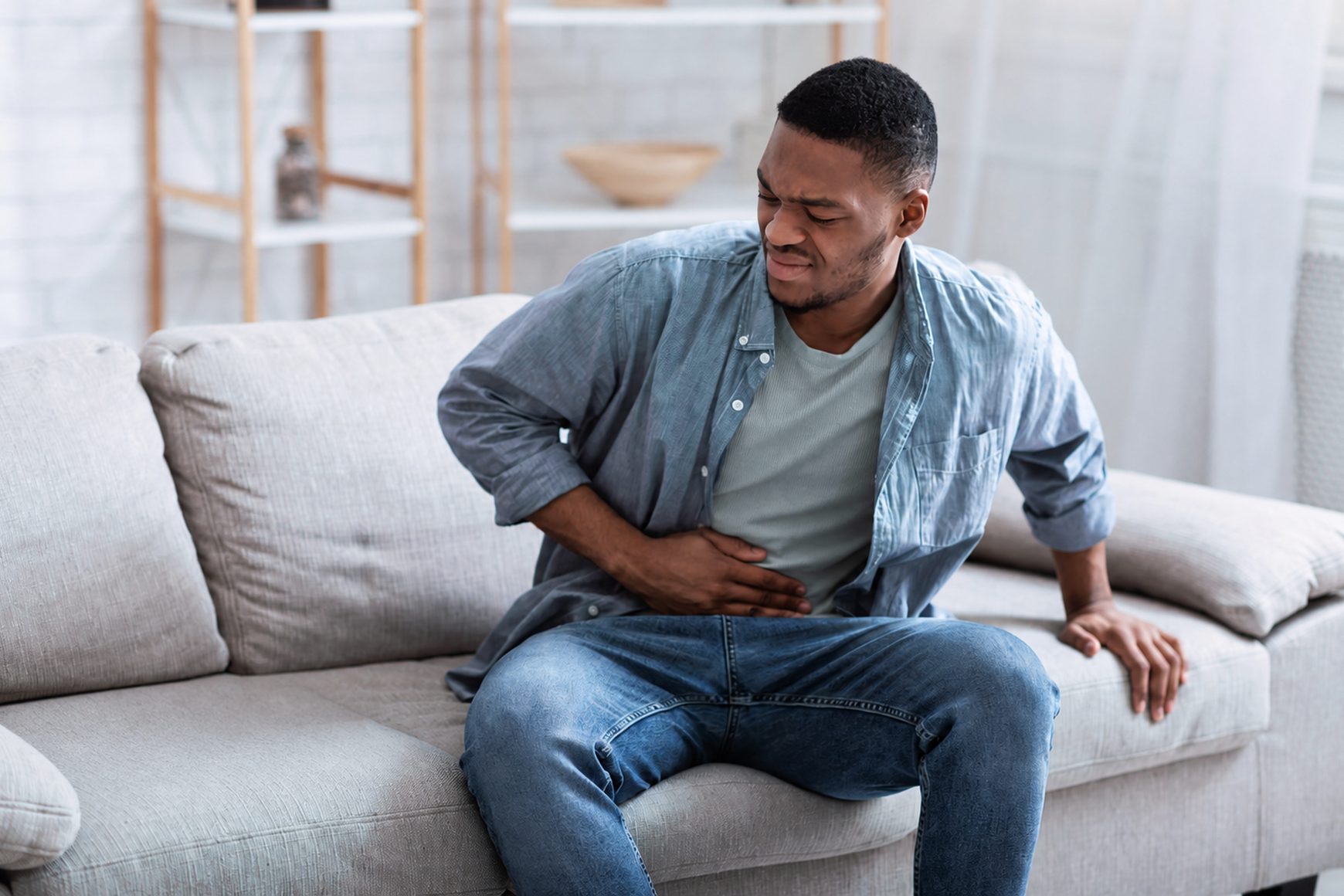 A young man sitting on a couch holding his abdomen in discomfort