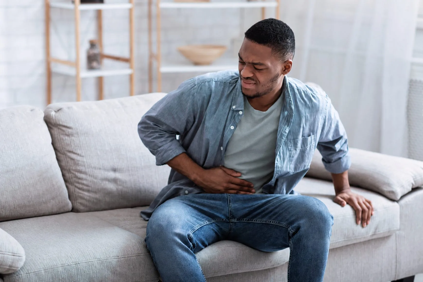 A young man sitting on a couch holding his abdomen in discomfort