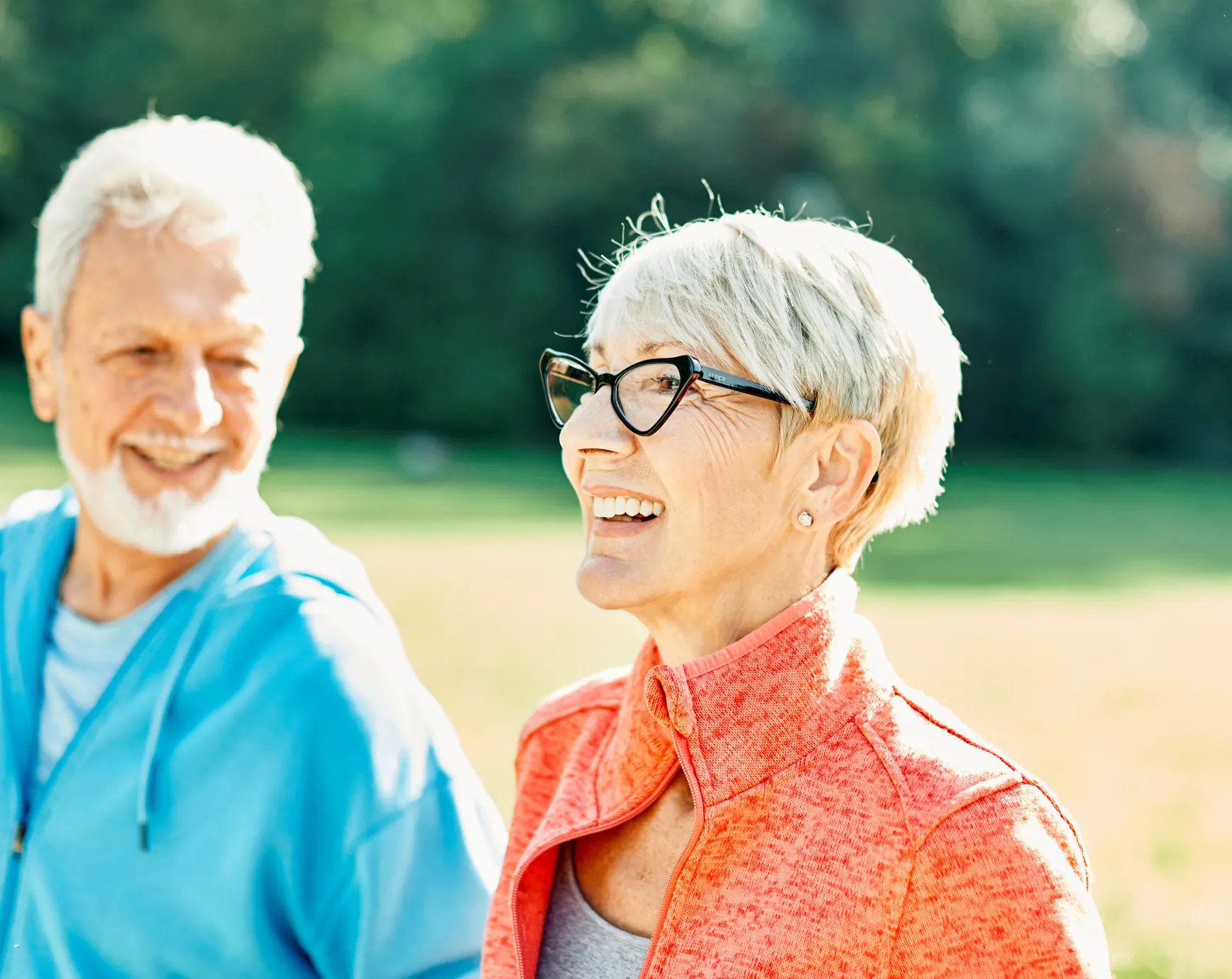 A smiling older woman in a coral jacket walking outdoors with her partner on a sunny day