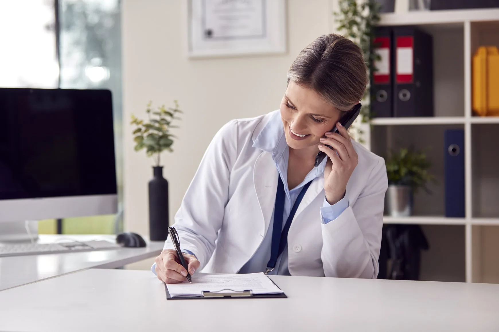 A smiling doctor in a white coat talking on the phone while writing notes on a clipboard at her desk