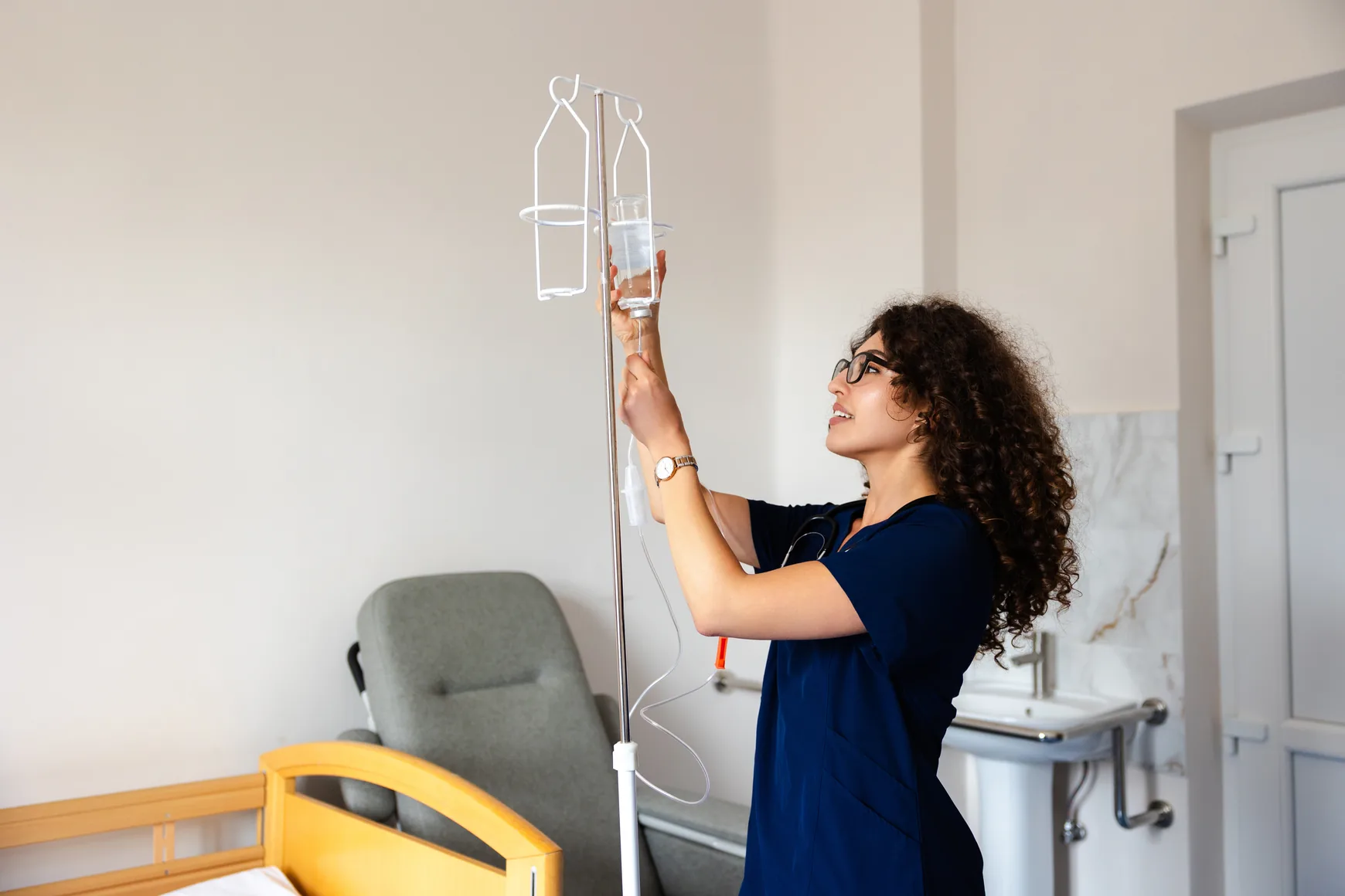 A nurse in scrubs adjusting an IV medication bag on a stand in a bright infusion room.