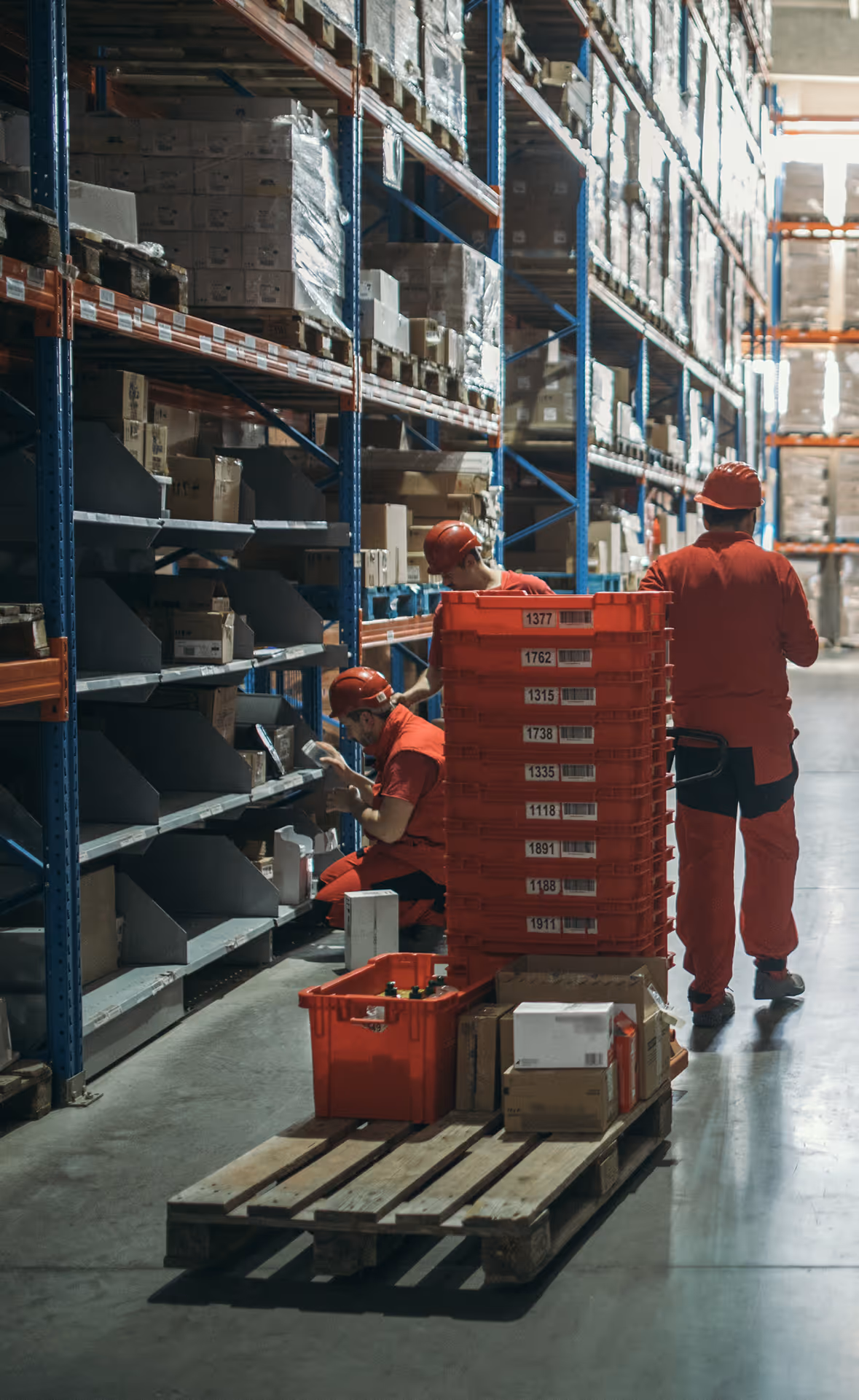 Warehouse workers in red uniforms and helmets organizing boxes on shelves and a pallet with red bins stacked in aisle.