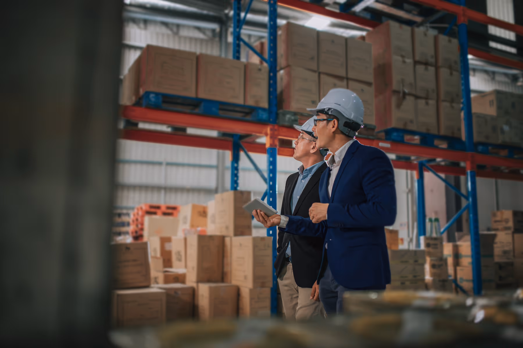 Two men wearing white hard hats and business attire inspecting stacked boxes in a warehouse.