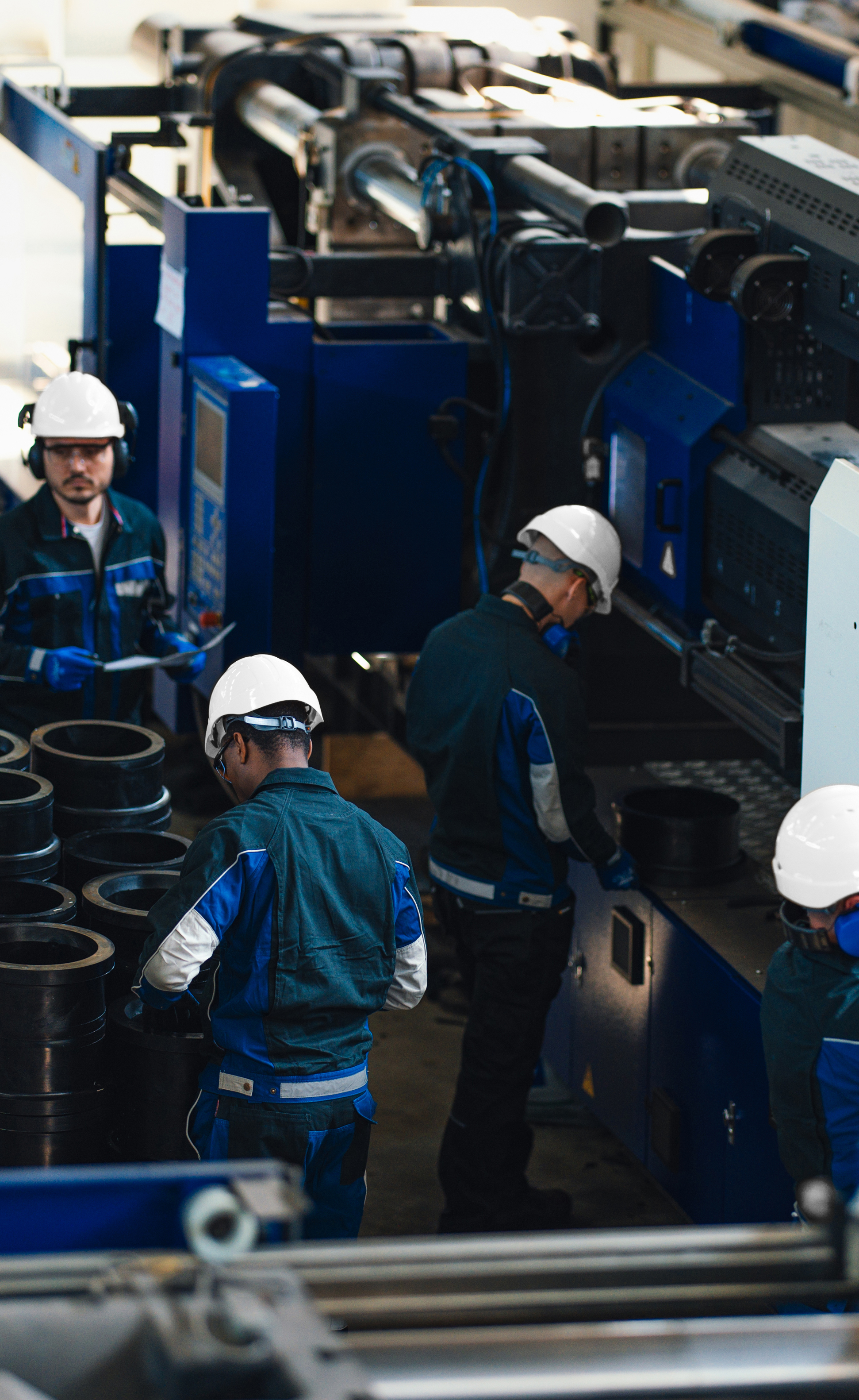 Factory workers wearing white helmets and blue uniforms working with industrial machinery and cylindrical metal parts.
