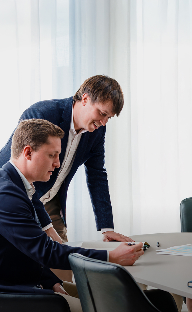 Two men in business attire reviewing documents at a table in an office with sheer white curtains.