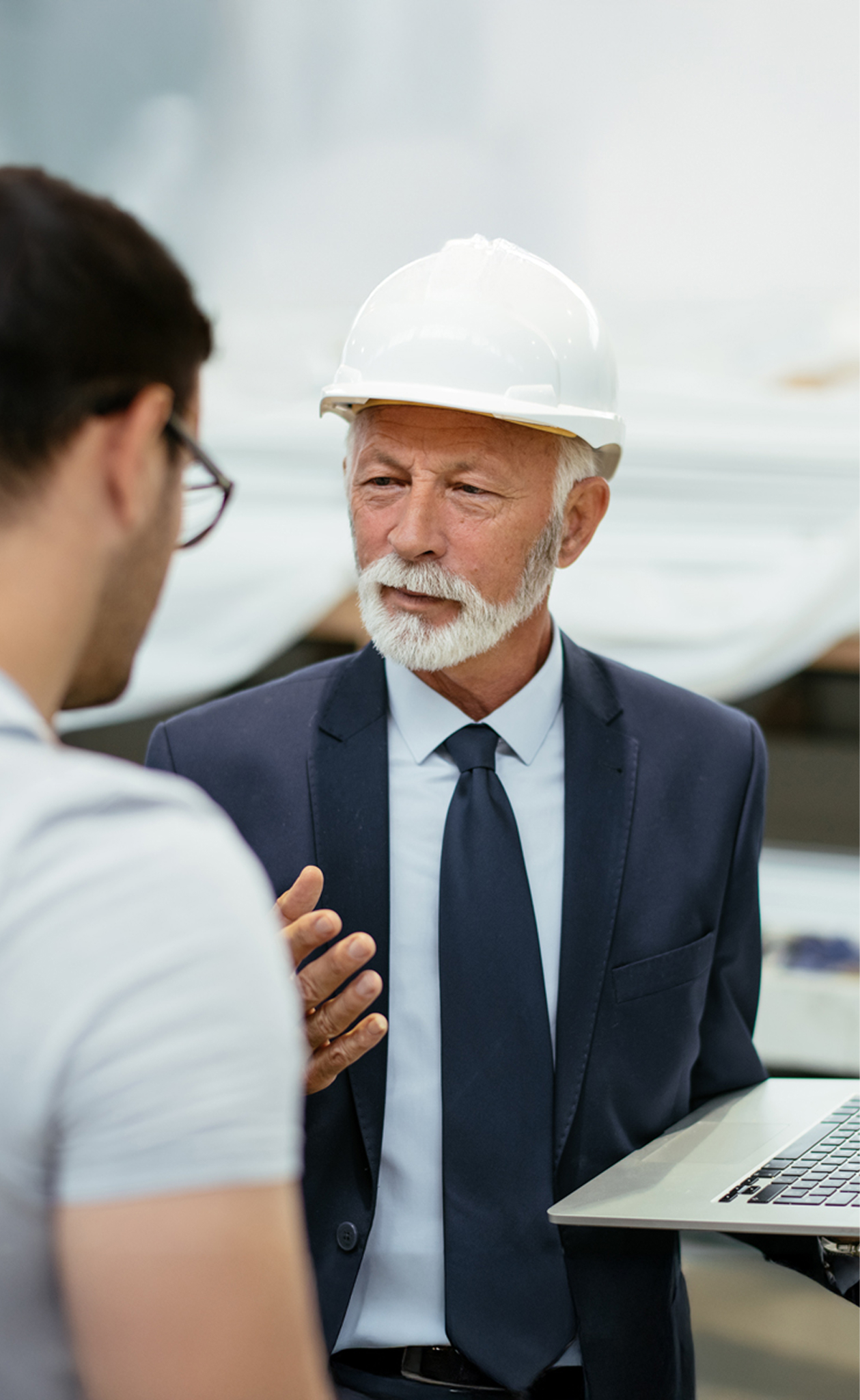 Senior man in a suit and white hard hat discussing while holding a laptop with a younger man.