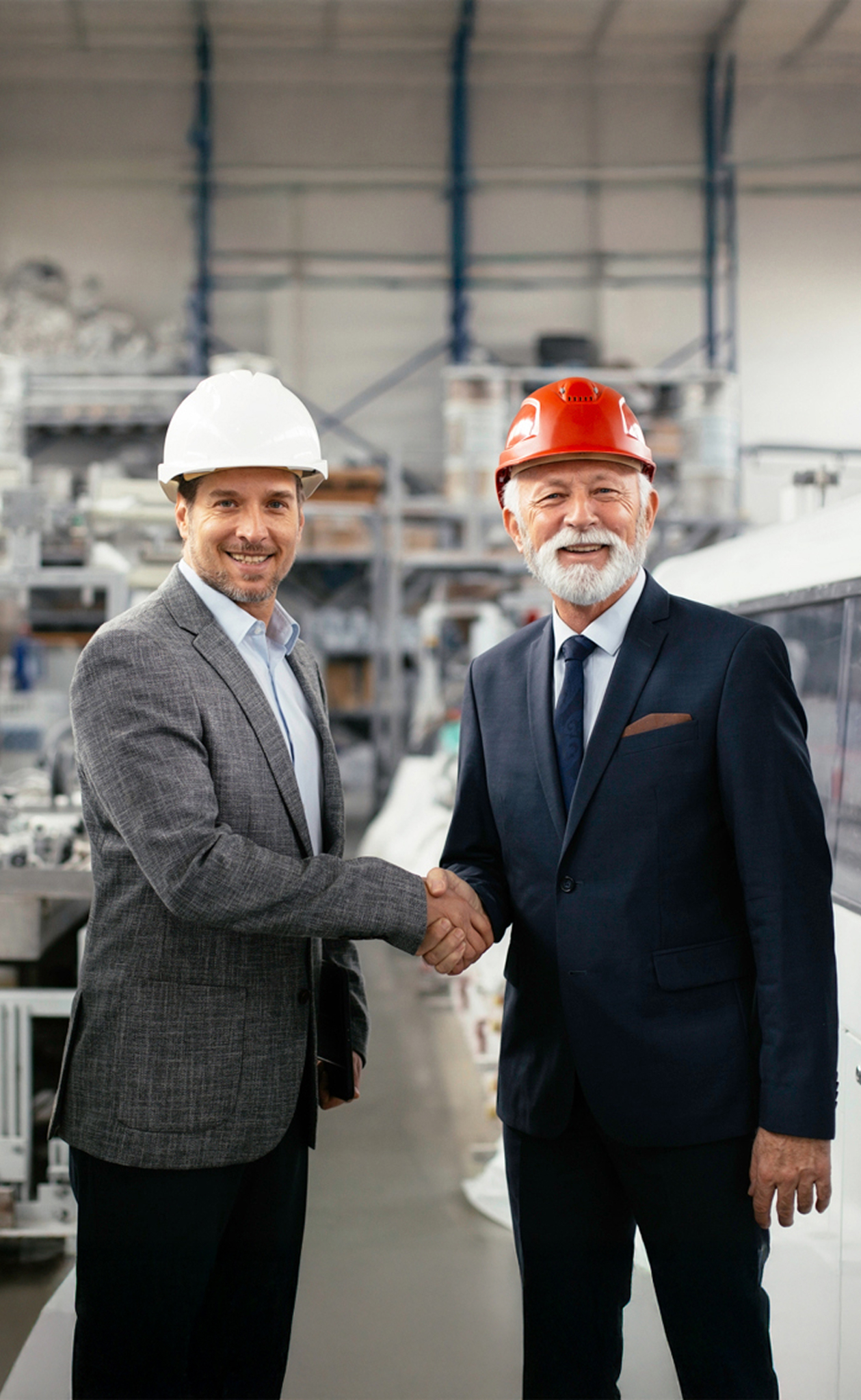 Two male professionals wearing safety helmets shaking hands in an industrial warehouse.