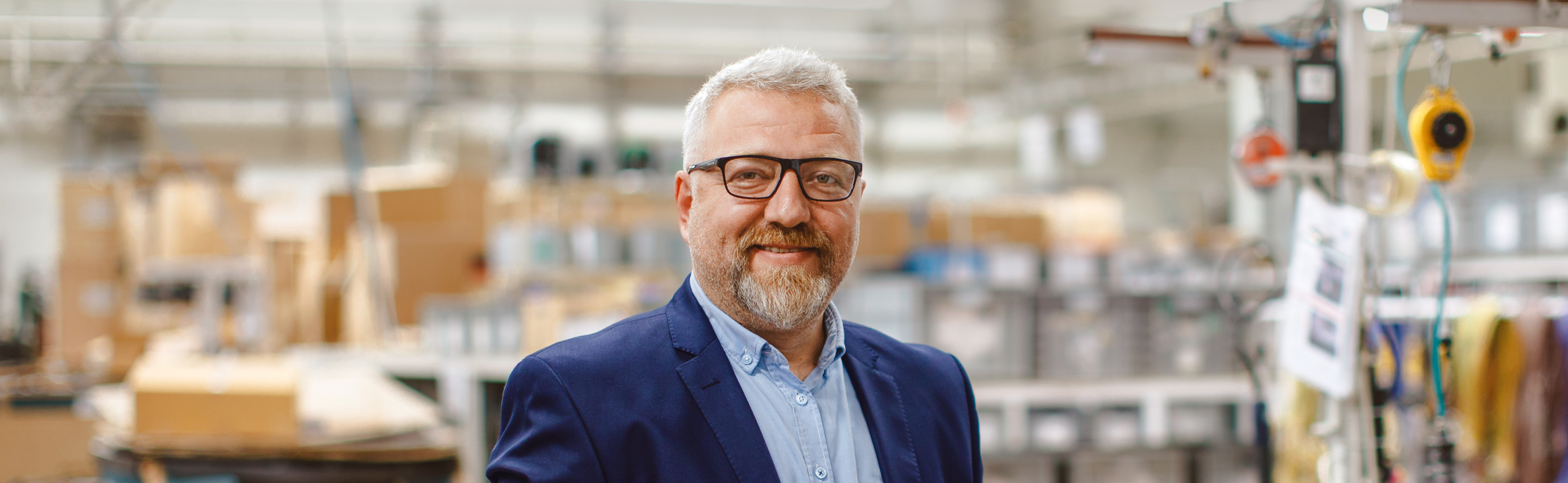 Smiling middle-aged man with gray hair and beard wearing glasses, a blue blazer, and a light blue shirt in an industrial warehouse.
